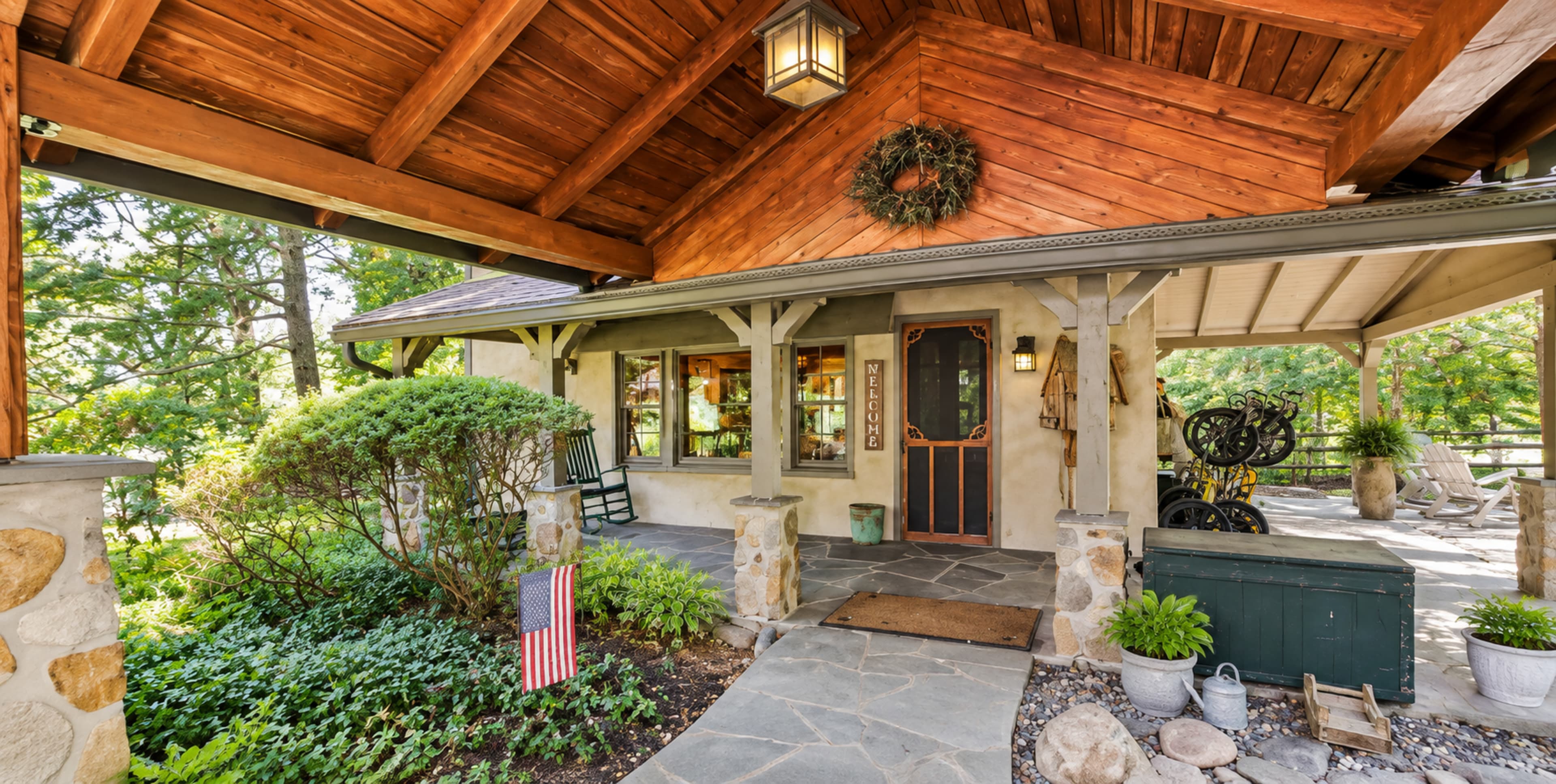 The image shows a rustic house entrance with a wooden overhang, stone pathway, and lush greenery, featuring an American flag near the door.