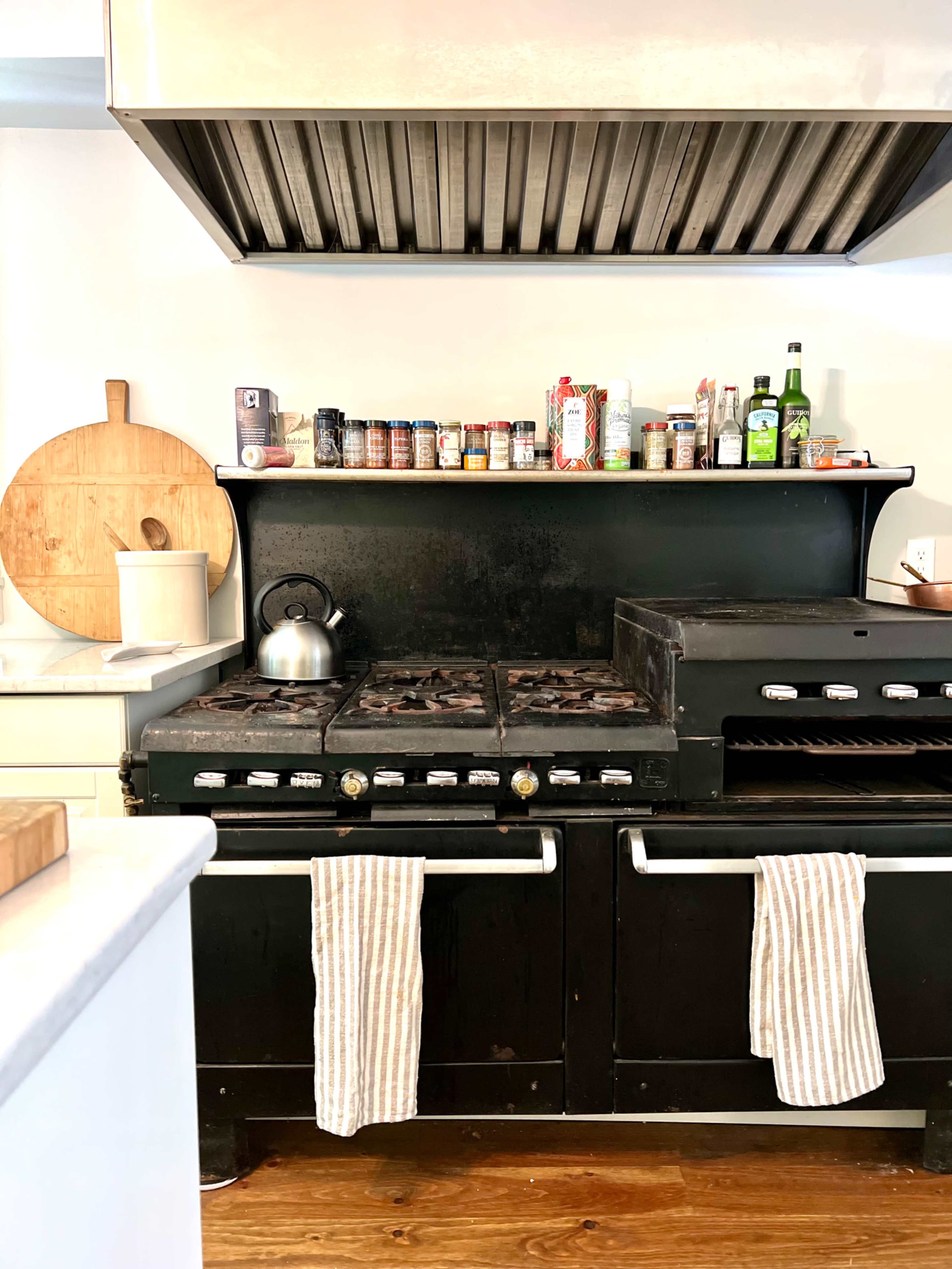 A black vintage stove with a kettle on it, surrounded by jars and cans on a shelf above, with striped tea towels hanging from the front.