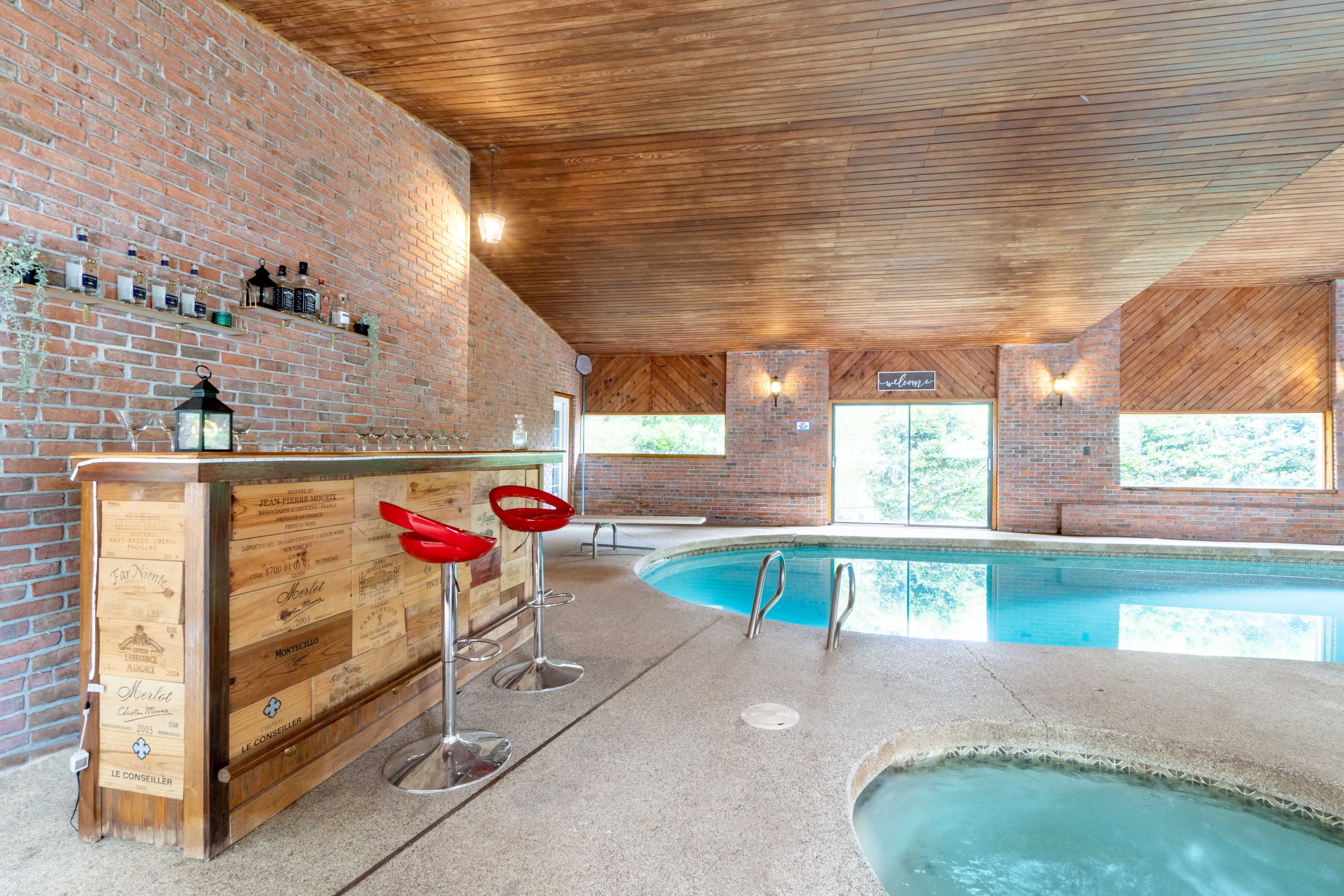 The image shows an indoor swimming area featuring a pool, a hot tub, and a bar with red stools against a brick wall.