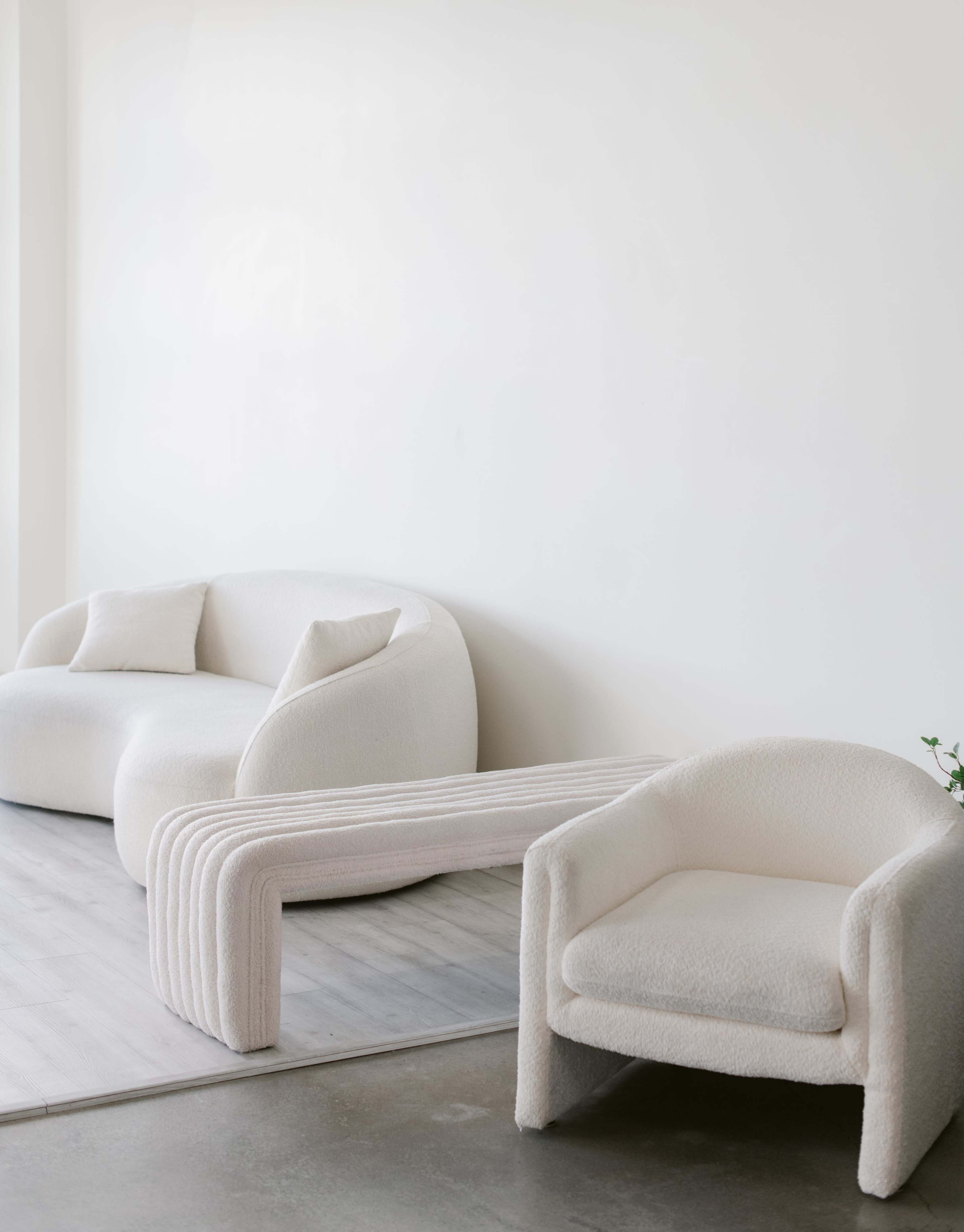 A minimalist seating arrangement with a white, plush sofa, a curvy white chair, and a sculptural coffee table against a light-colored wall and wooden floor.