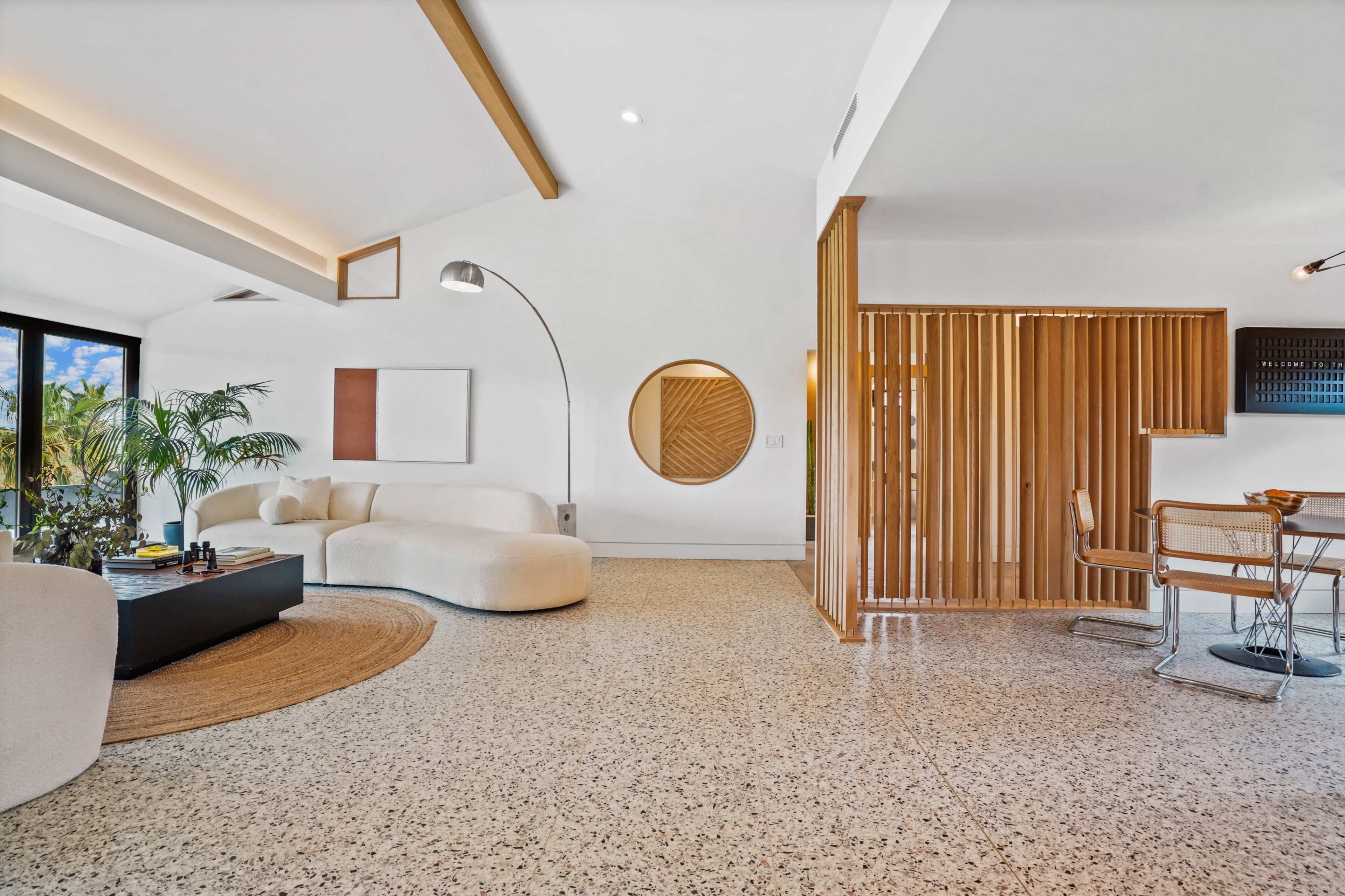 The image shows a modern living space with a curved white sofa, a round coffee table, and a textured circular area rug, complemented by wooden accents and a glass wall.
