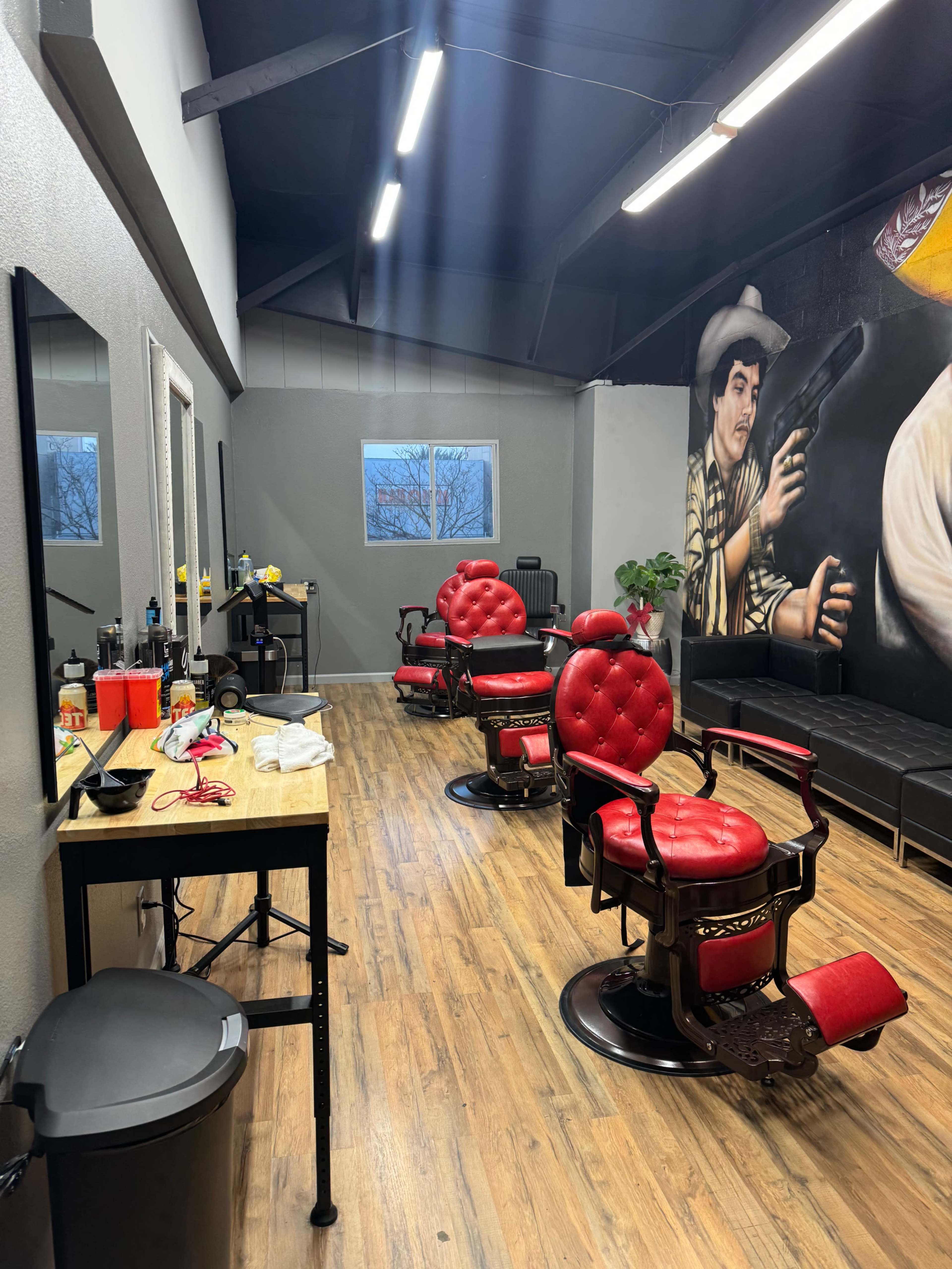The image shows a modern barbershop featuring two red barber chairs, a wooden work table, and large mirrors against gray walls, with a mural of a figure holding a gun in the background.
