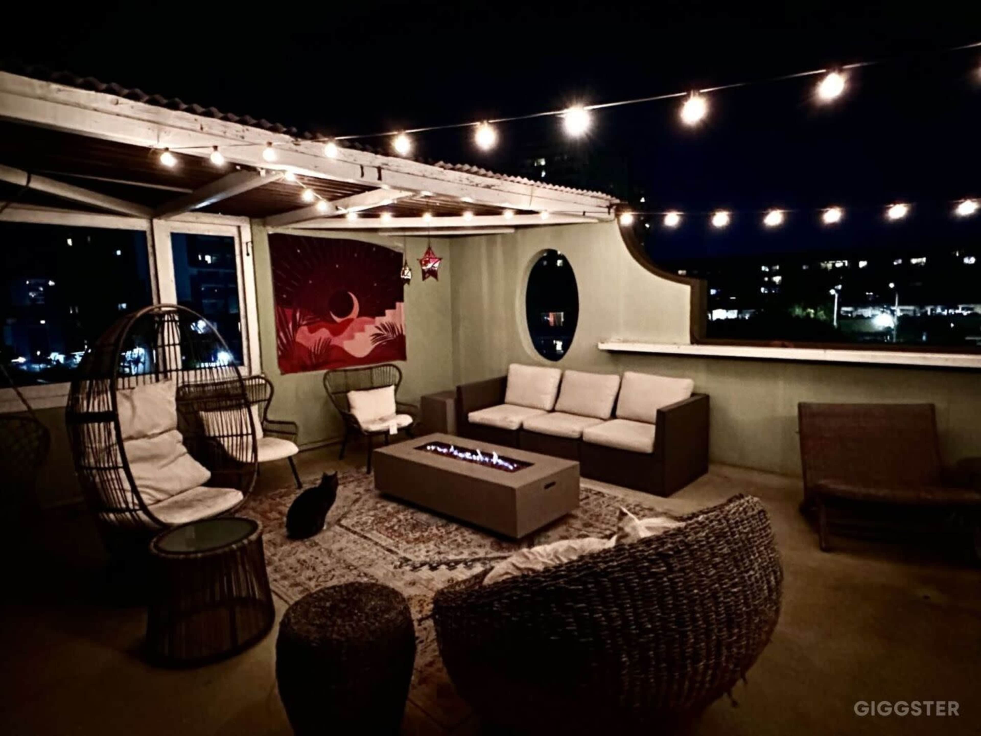 A rooftop patio features a fire pit surrounded by seating, decorative string lights overhead, and a cat resting on the floor.