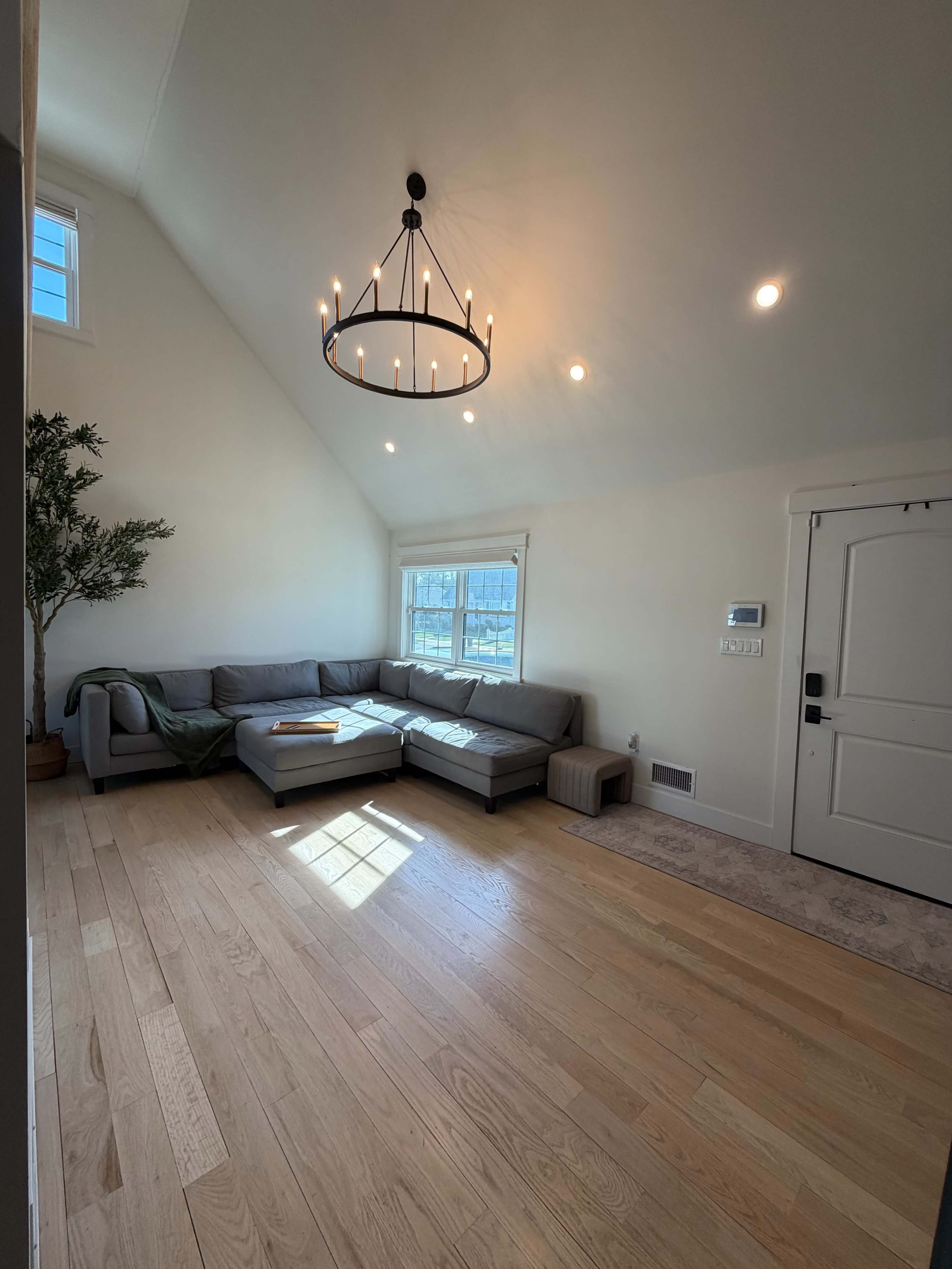 The image shows a well-lit living room featuring a gray sectional sofa, a chandelier, large windows, and light wooden flooring.