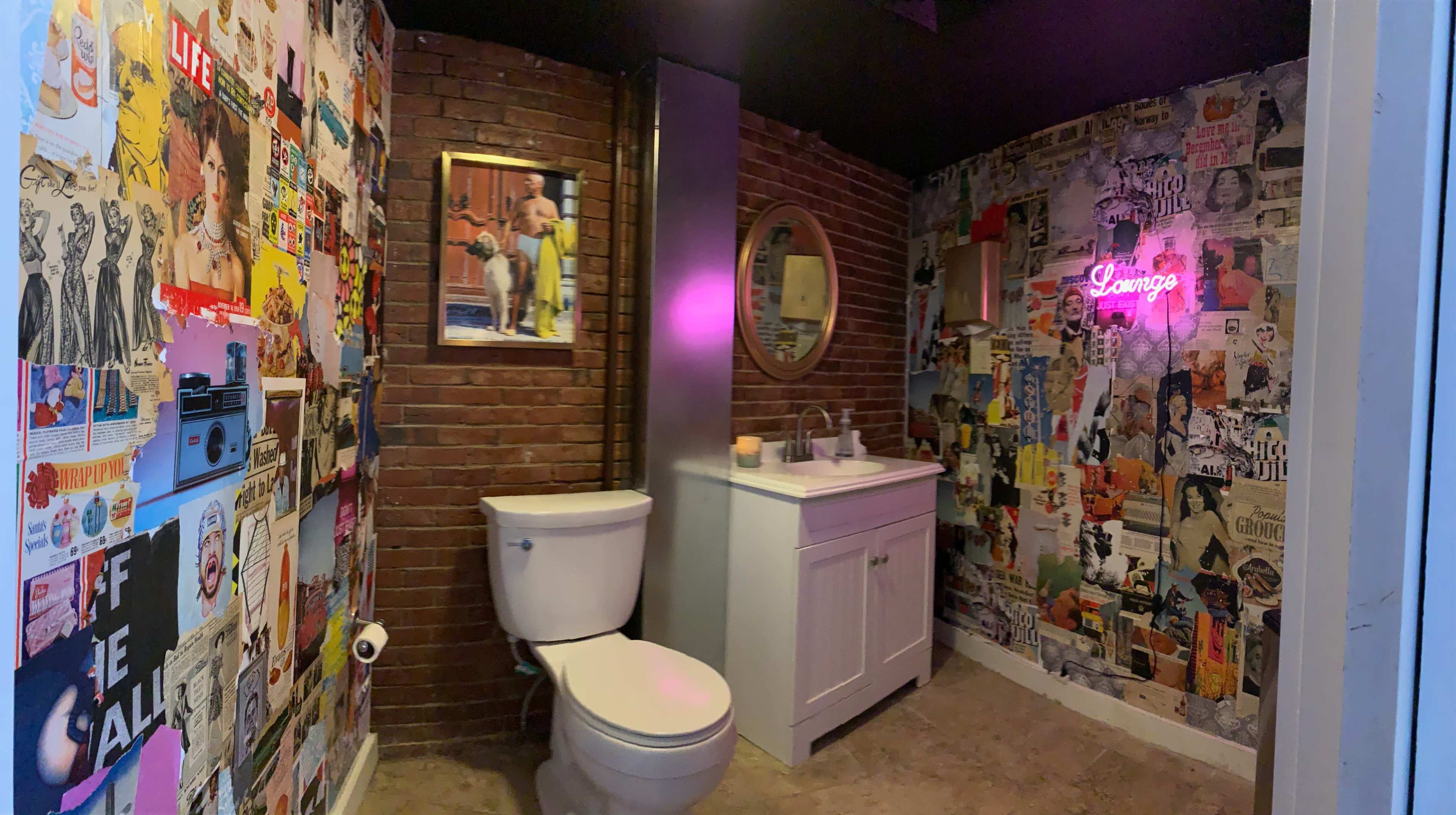 A bathroom features a white toilet and vanity against walls adorned with colorful posters and a neon sign.