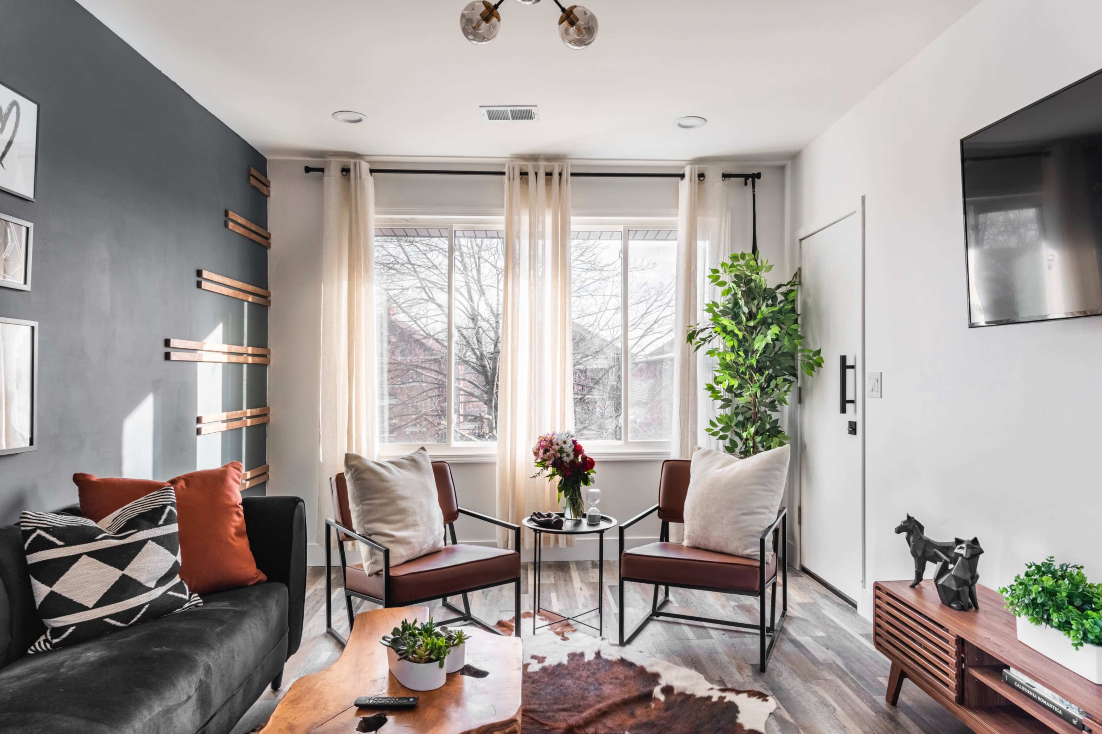 A modern living room featuring a dark accent wall, two chairs with cushions, a coffee table, and large windows with sheer curtains allowing natural light to fill the space.