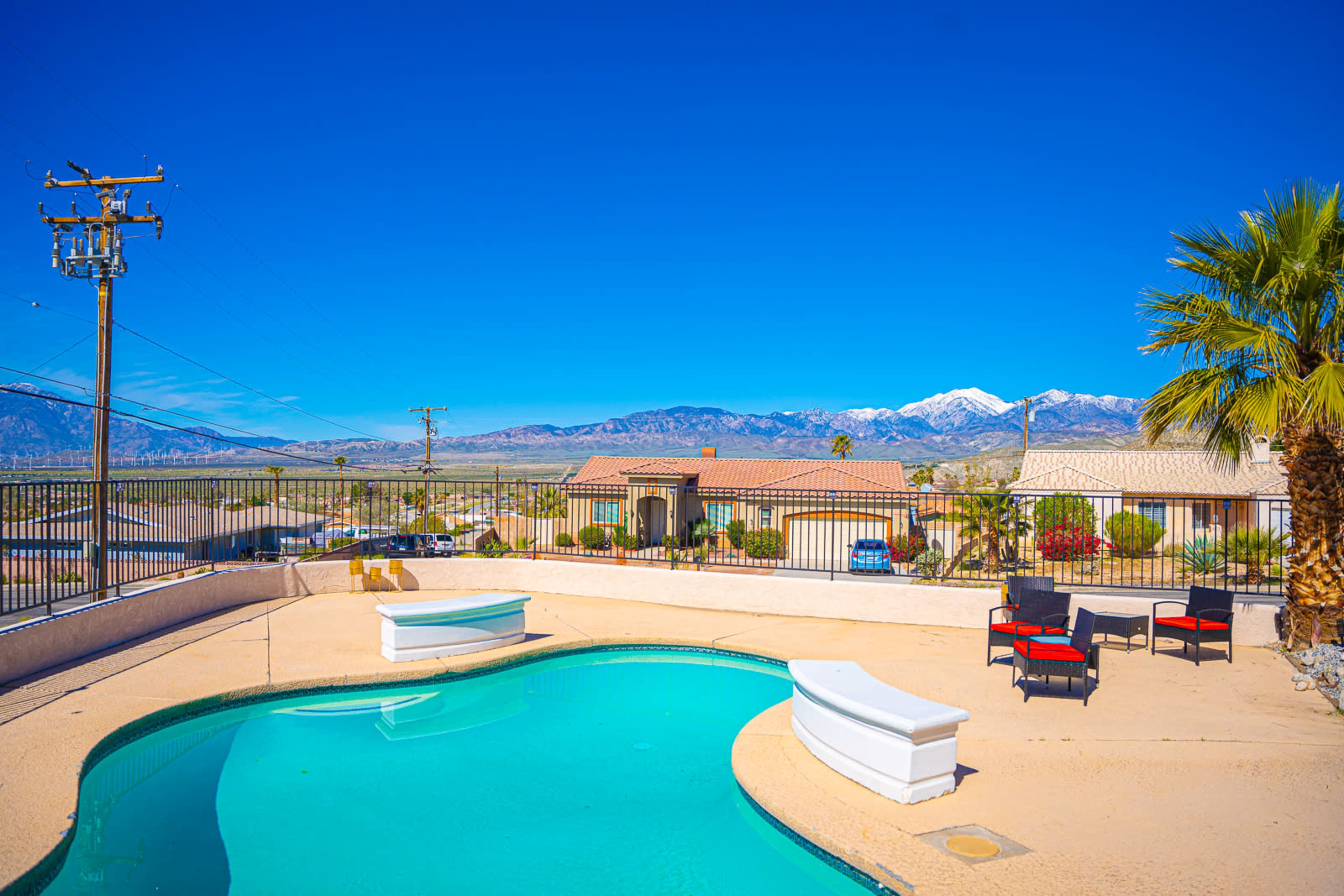 A swimming pool surrounded by lounge chairs is situated on a patio, with mountains and blue skies in the background.