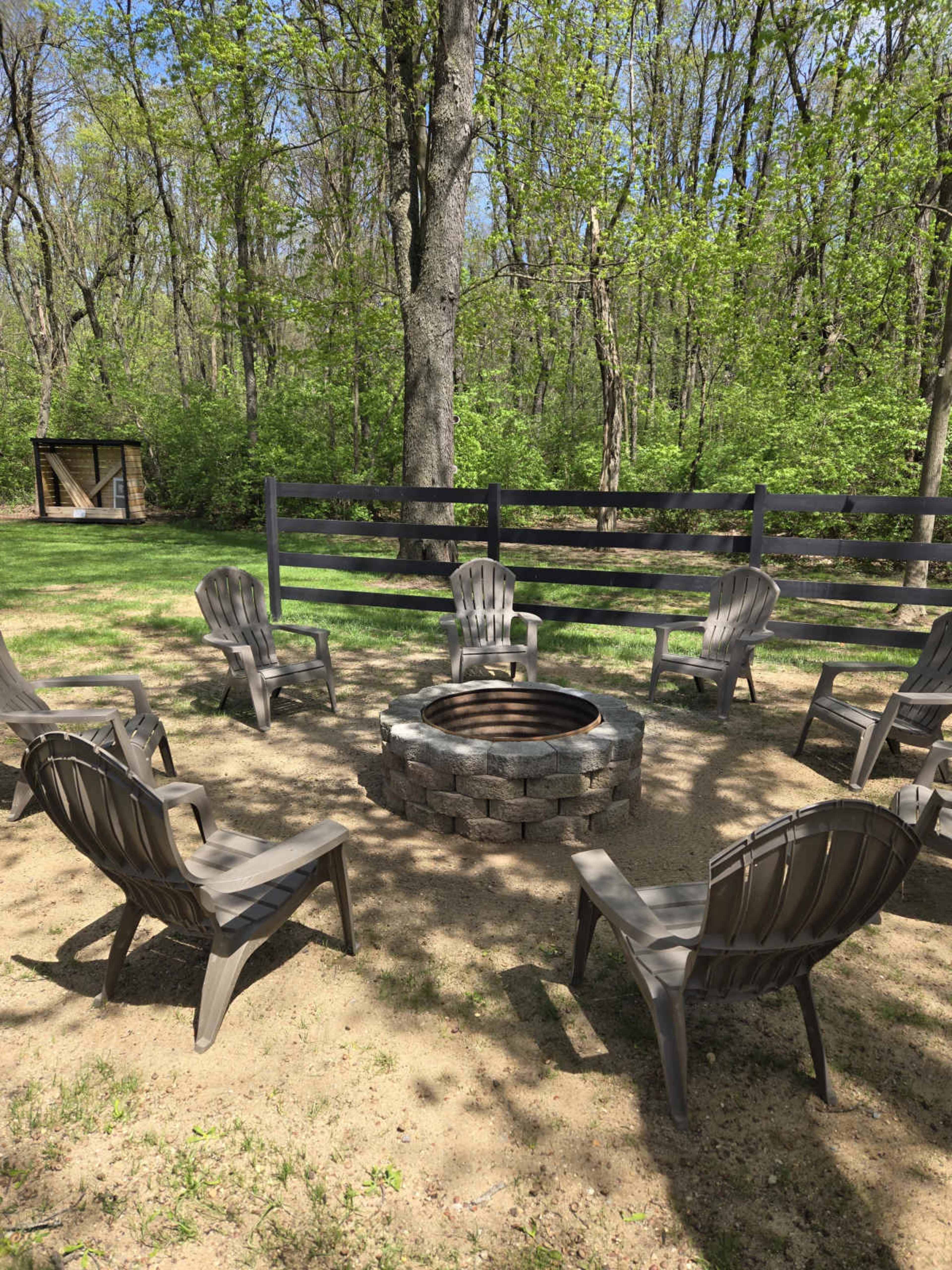 A stone fire pit surrounded by several gray Adirondack chairs is set in a wooded area with green foliage.