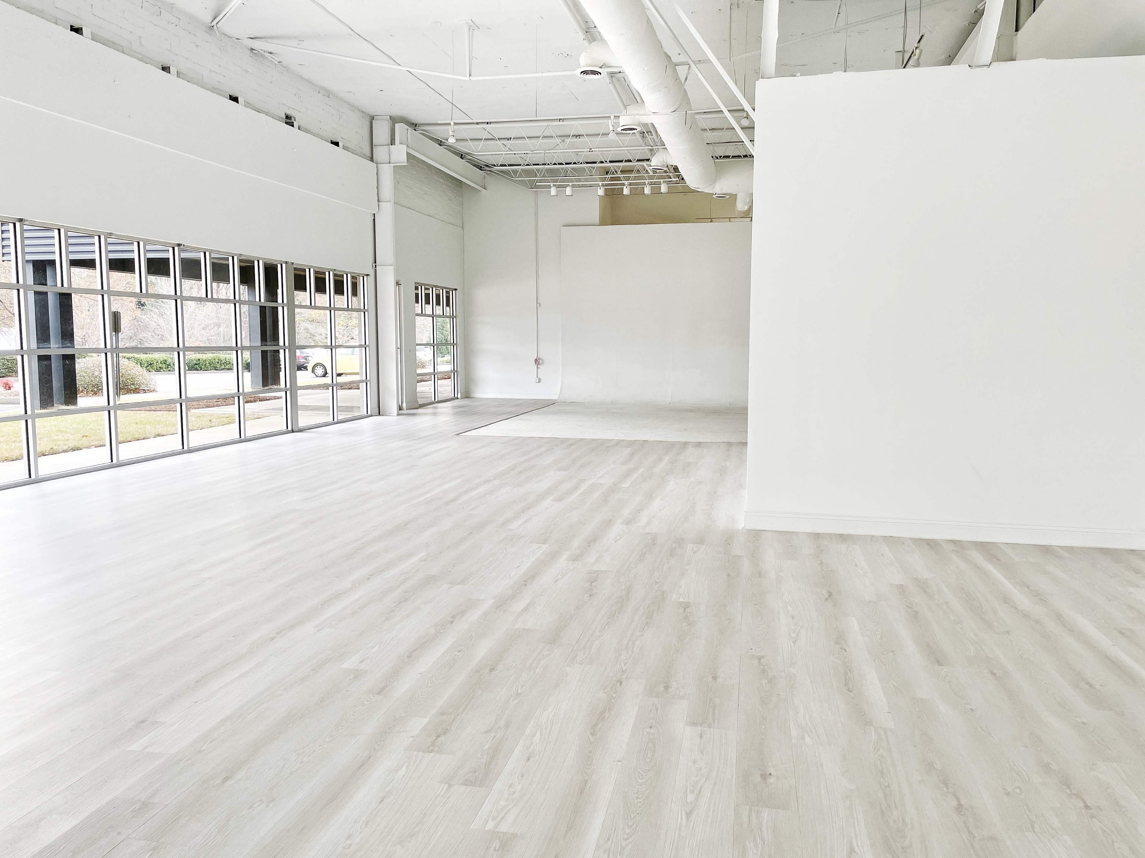 A large, empty interior space with light-colored flooring, large windows on one side, and a plain white wall at the back.