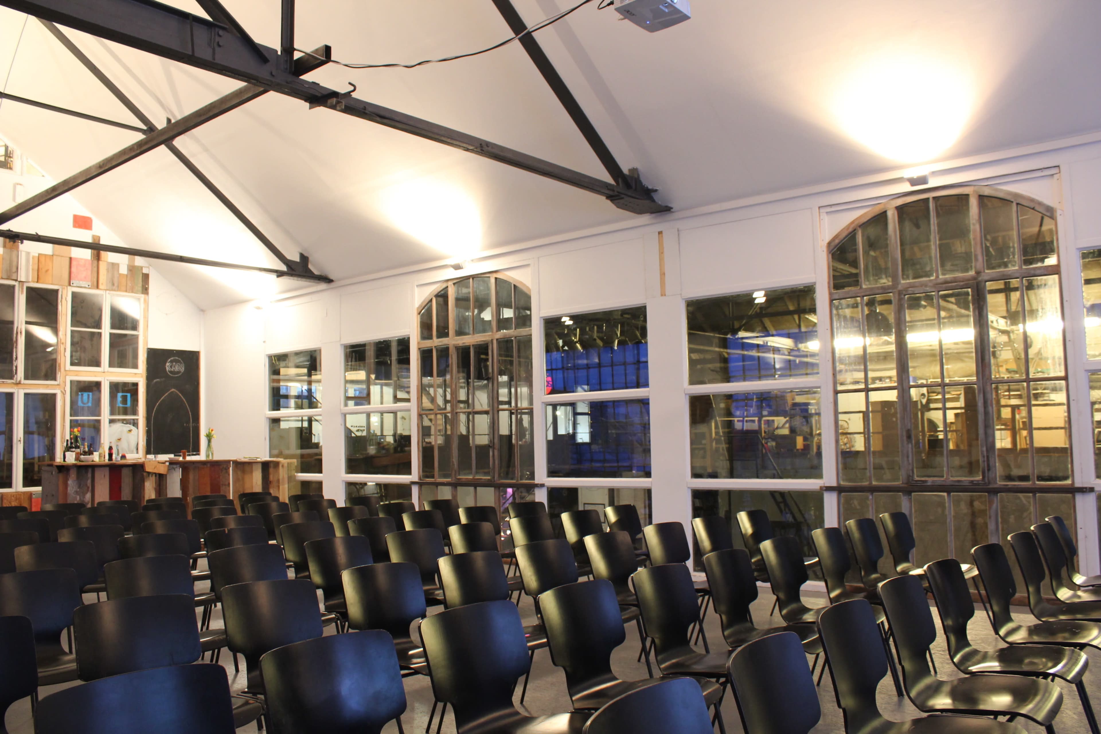 The image shows a spacious room with rows of black chairs arranged in front of large windows and a bar area in the background.