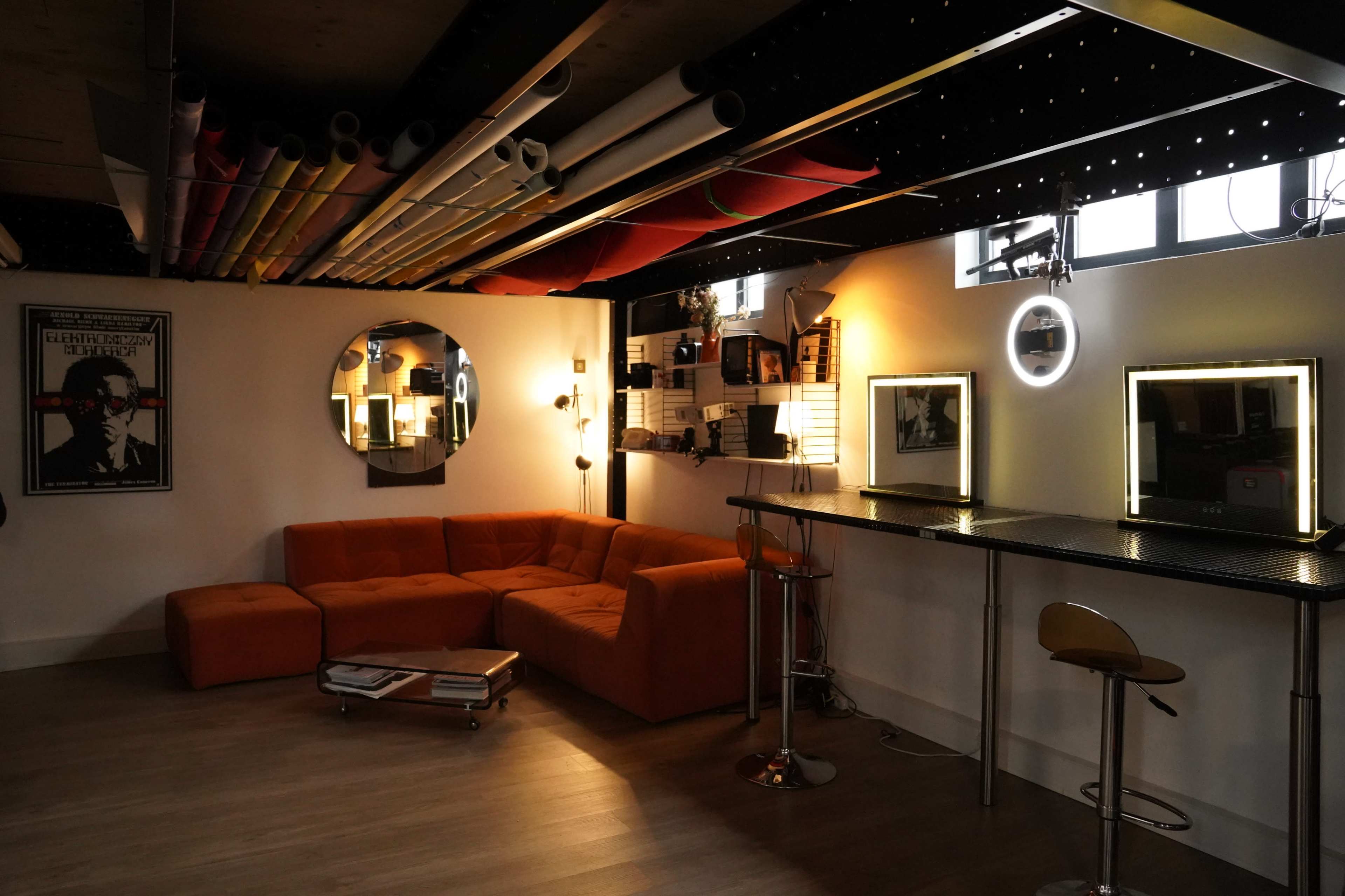 A modern lounge area features an orange sectional sofa, a bar with stools, and large mirrors under a ceiling adorned with colorful pipes.