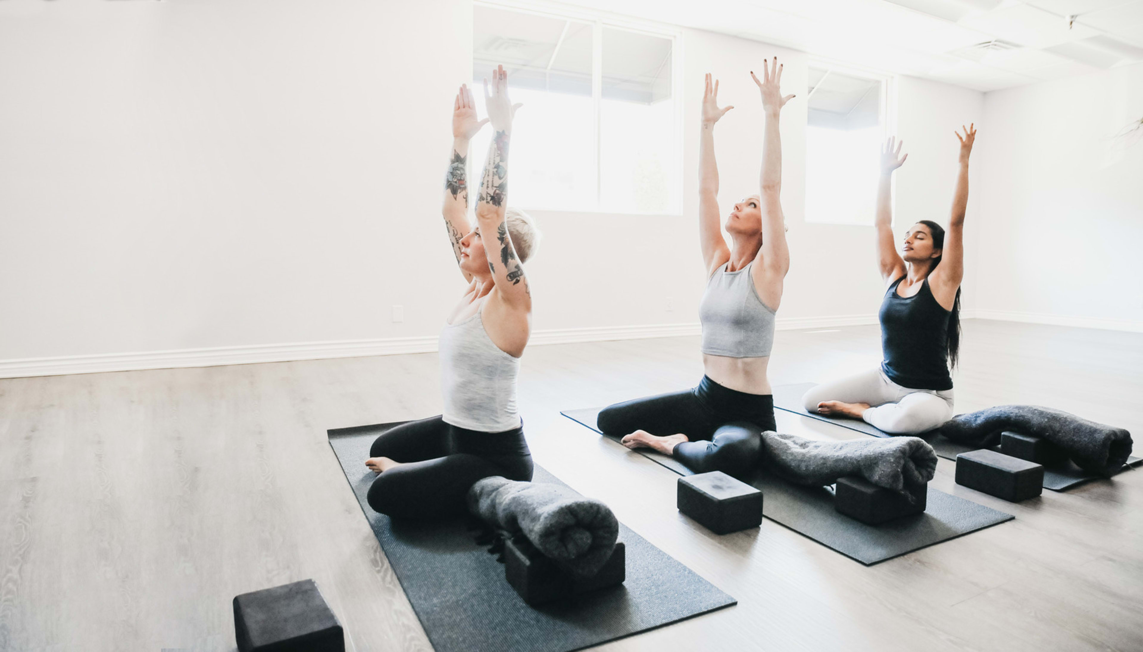 Three women practice yoga in a bright studio, each stretching with their arms raised while seated on mats.