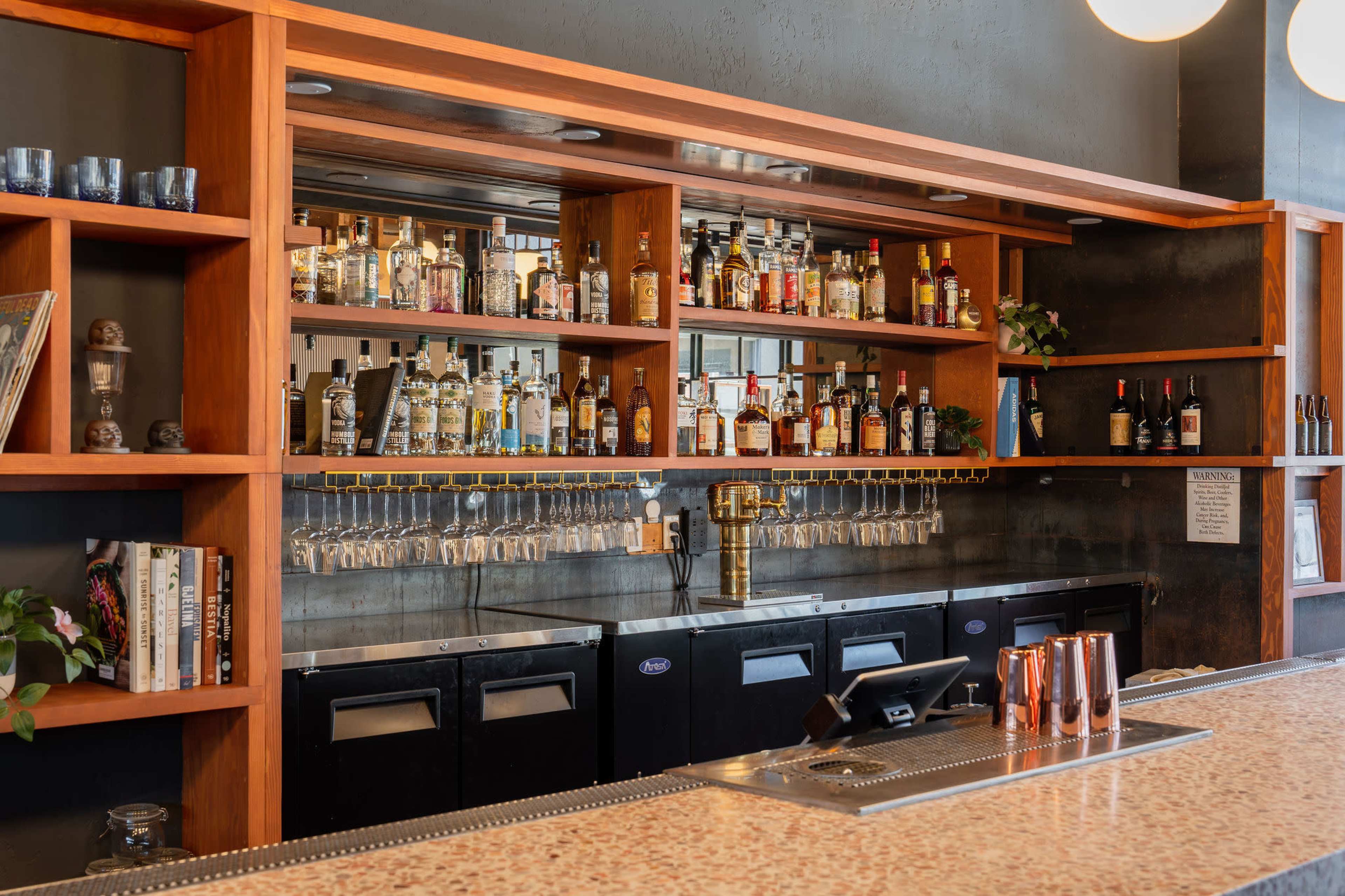 A bar with a wooden shelf filled with various bottles of liquor, glassware hanging beneath, and a polished countertop in front.