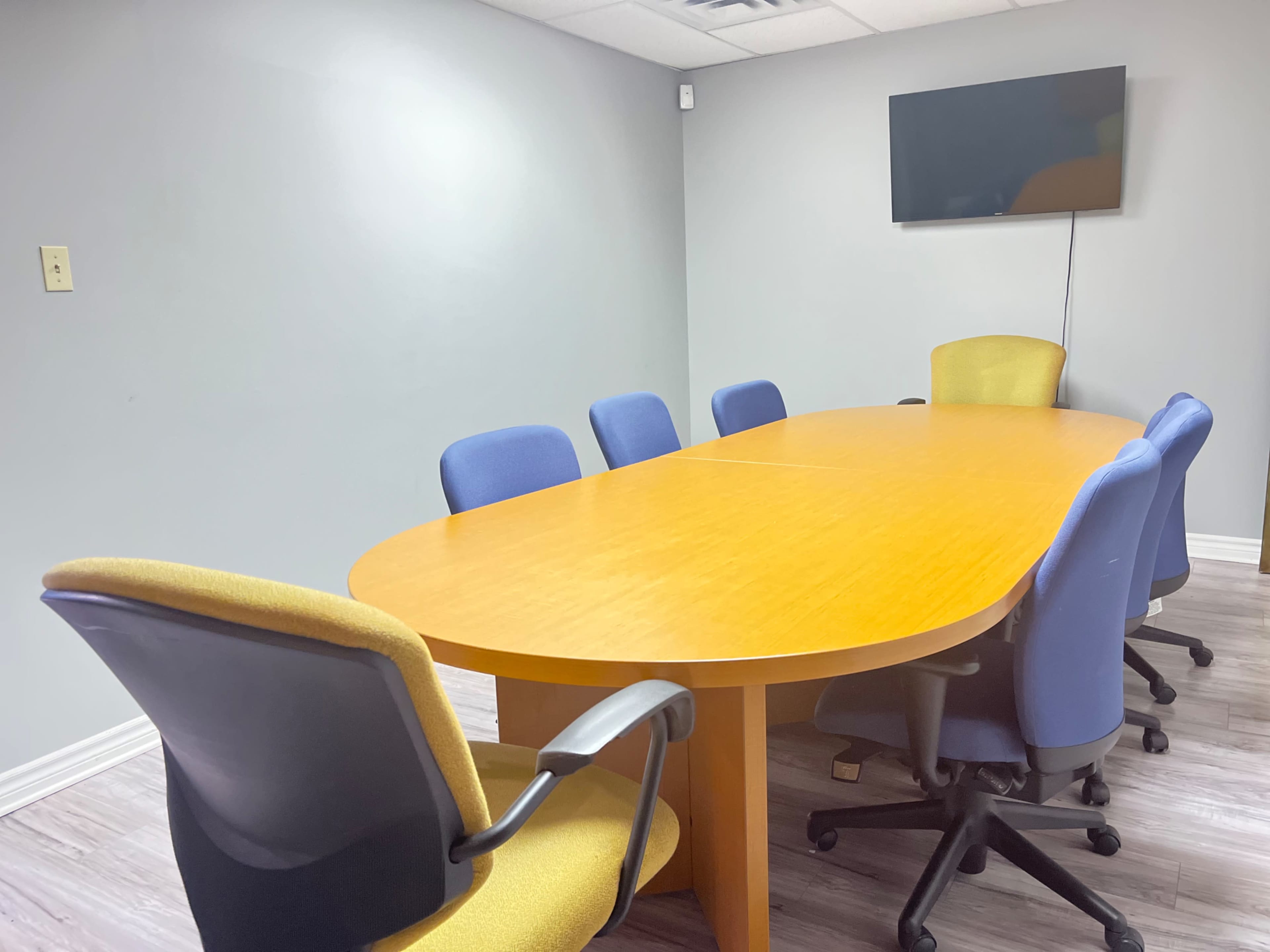 A conference room features a large oval wooden table surrounded by eight office chairs, with a wall-mounted television in the background.