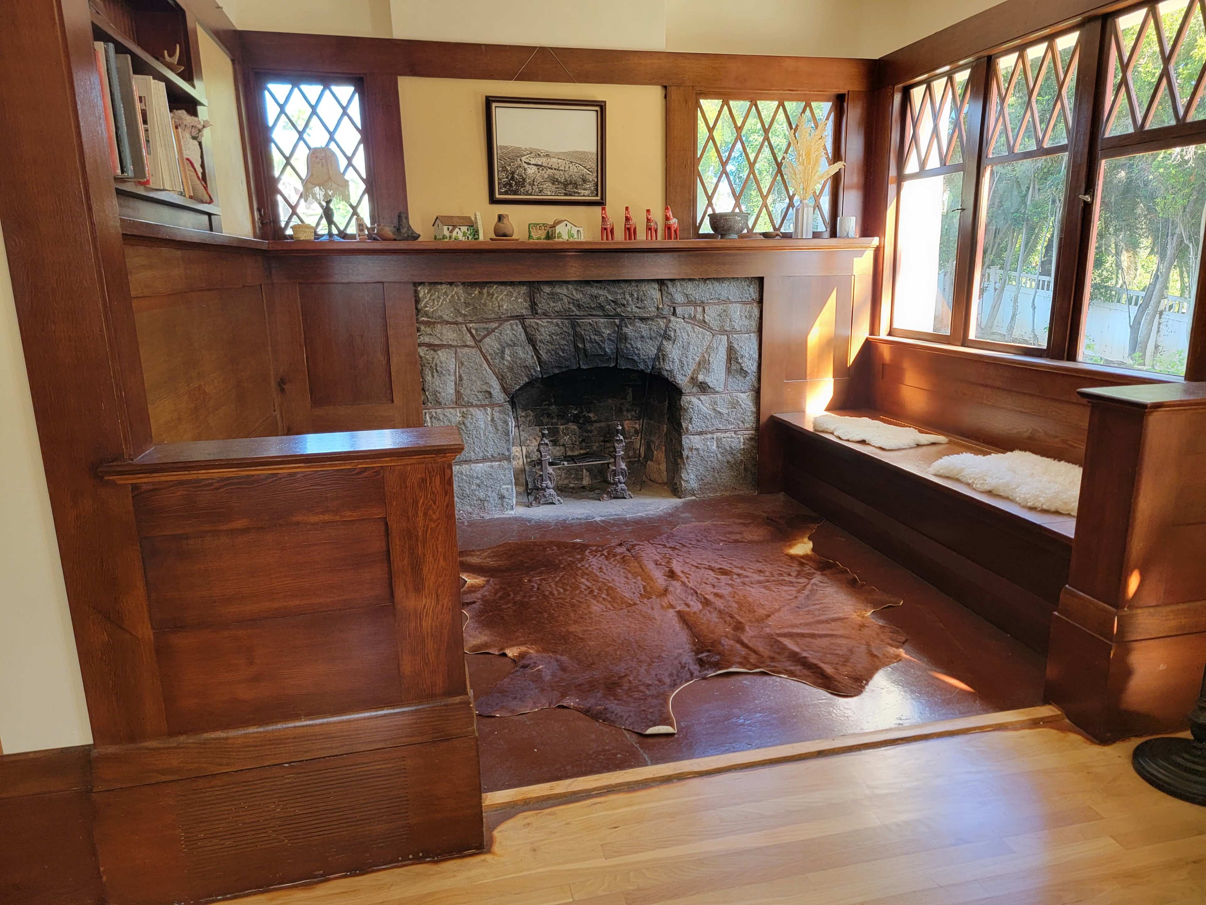 The image shows a cozy room featuring a stone fireplace, wooden paneling, and a bench with a soft fur covering, complemented by large windows.