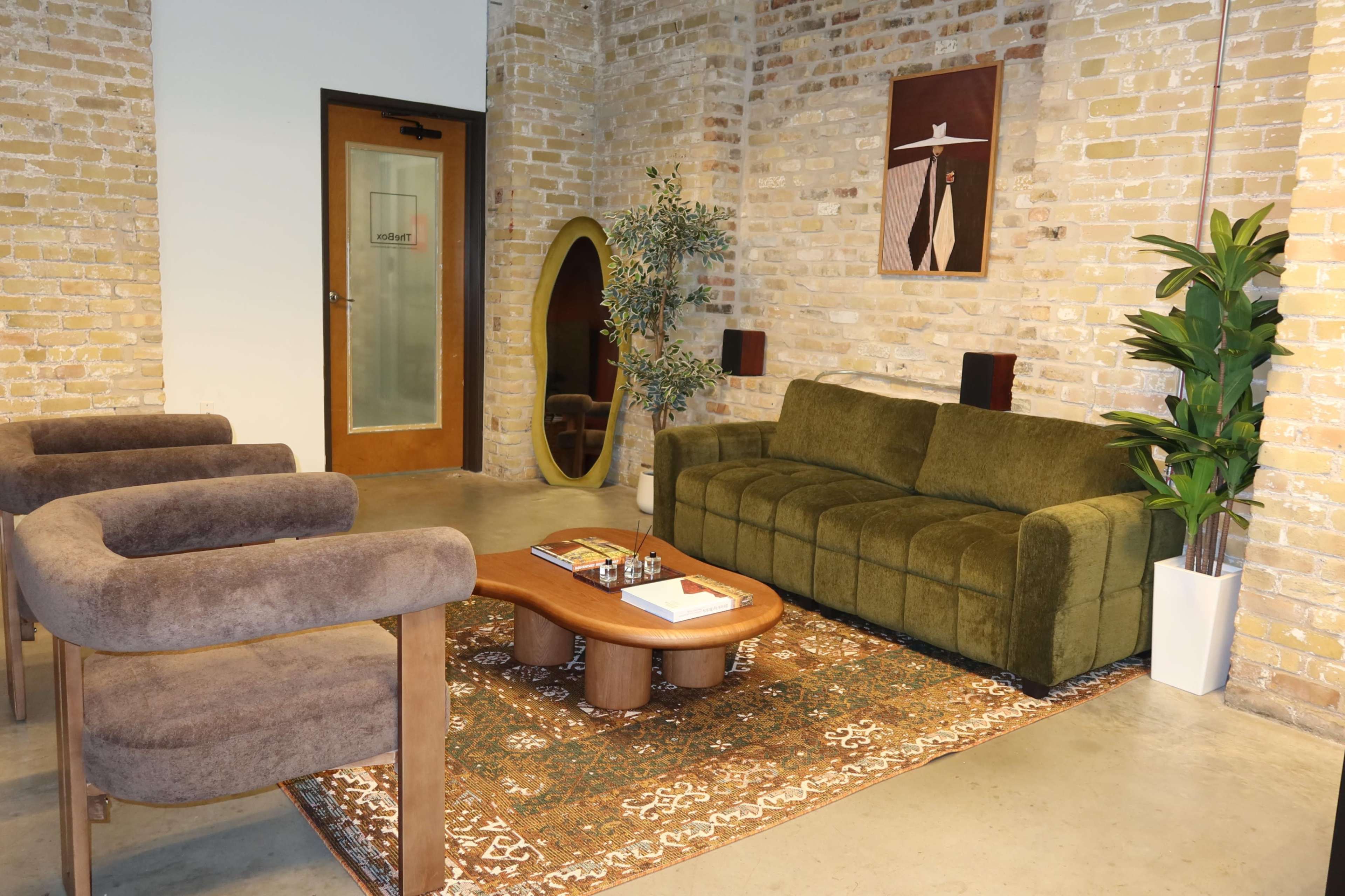 The image shows a modern lounge area with a green sofa, two brown armchairs, a circular mirror, a wooden coffee table, and decorative plants, all set against a brick wall.