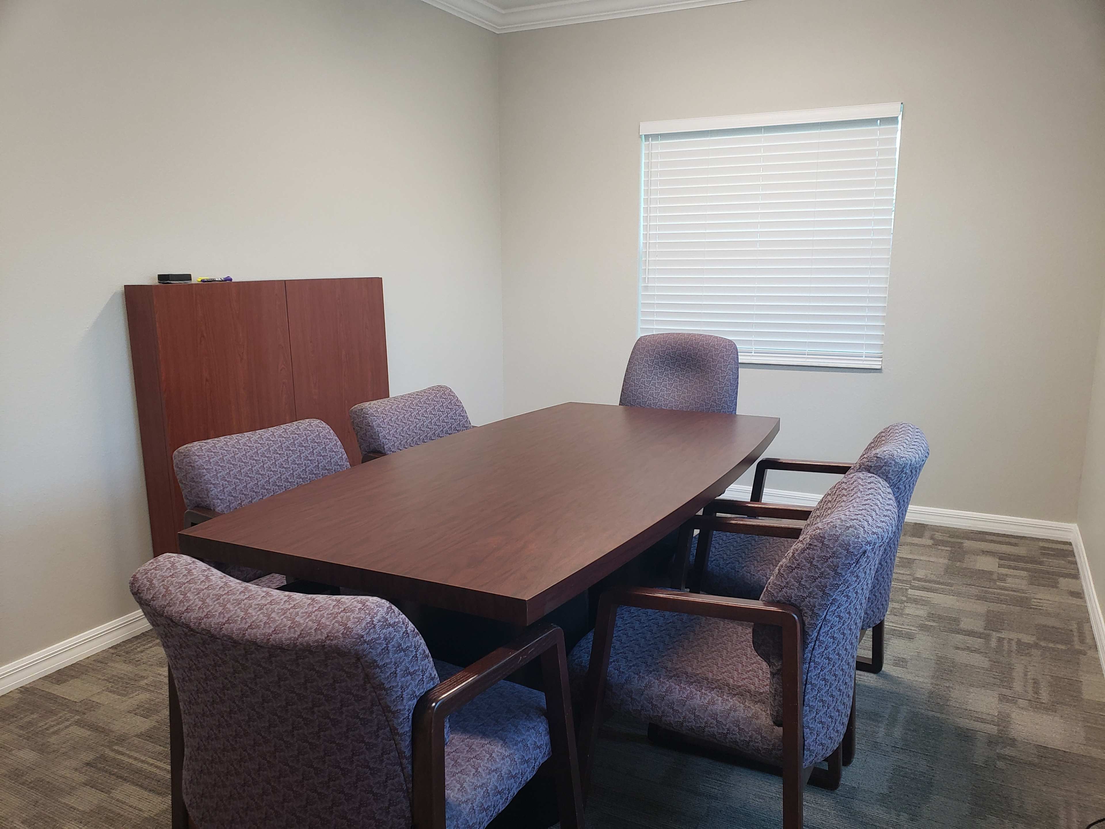 A conference room contains a large table surrounded by six upholstered chairs and a cabinet against the wall.