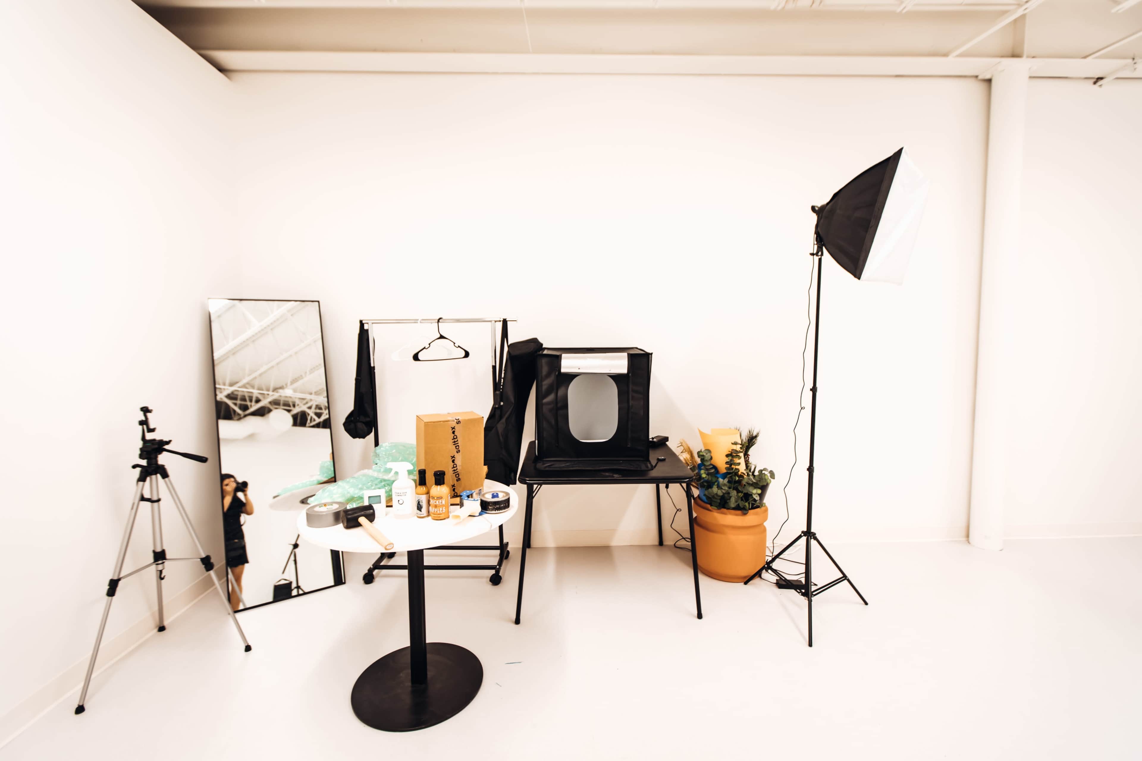 A small studio space features a table with various beauty products, a tripod, a mirror, a chair with a portable backdrop, and a potted plant.