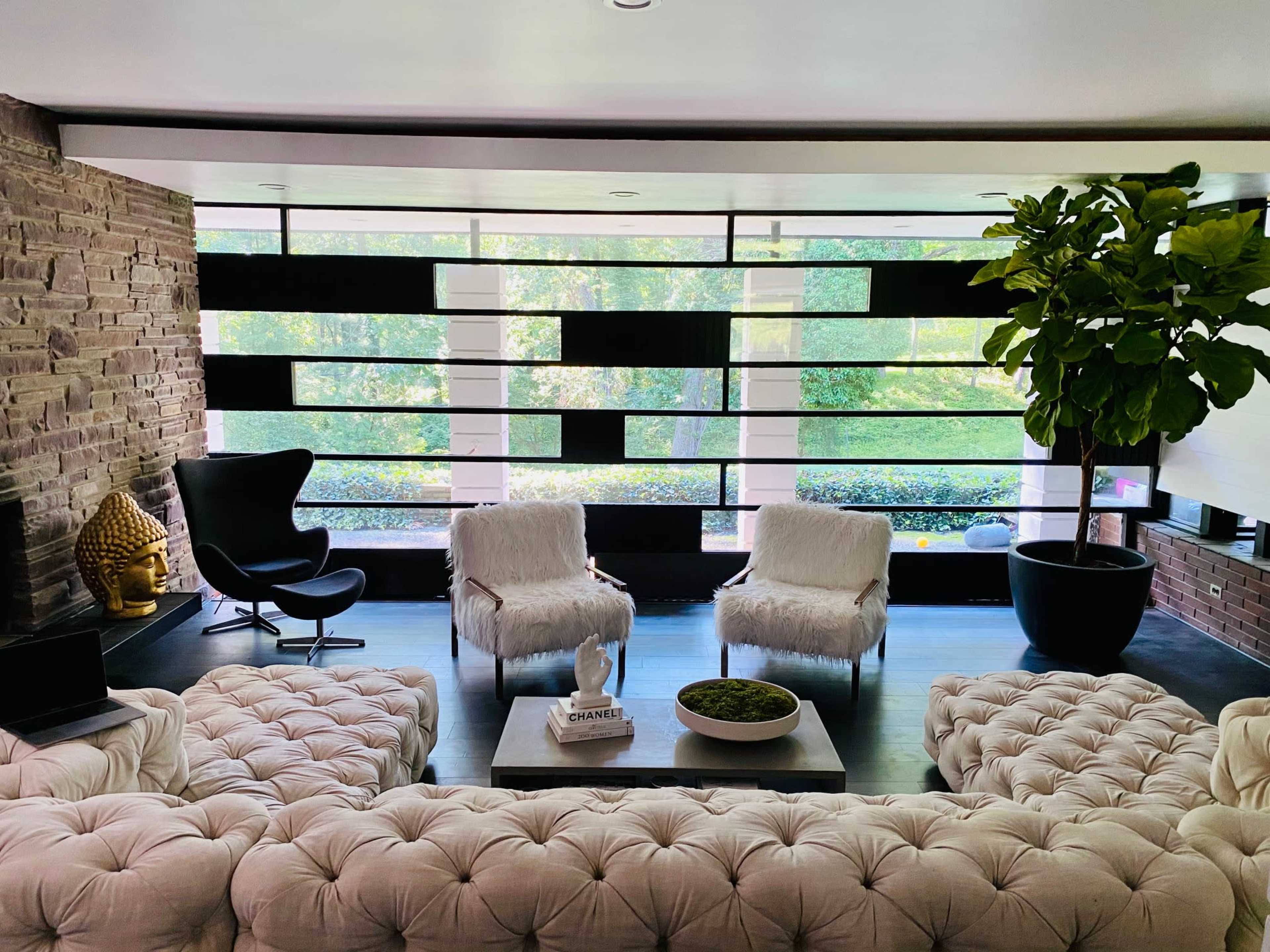 A modern living room with tufted seating, a glass wall providing views of greenery, and decorative elements including a plant and a Buddha statue.