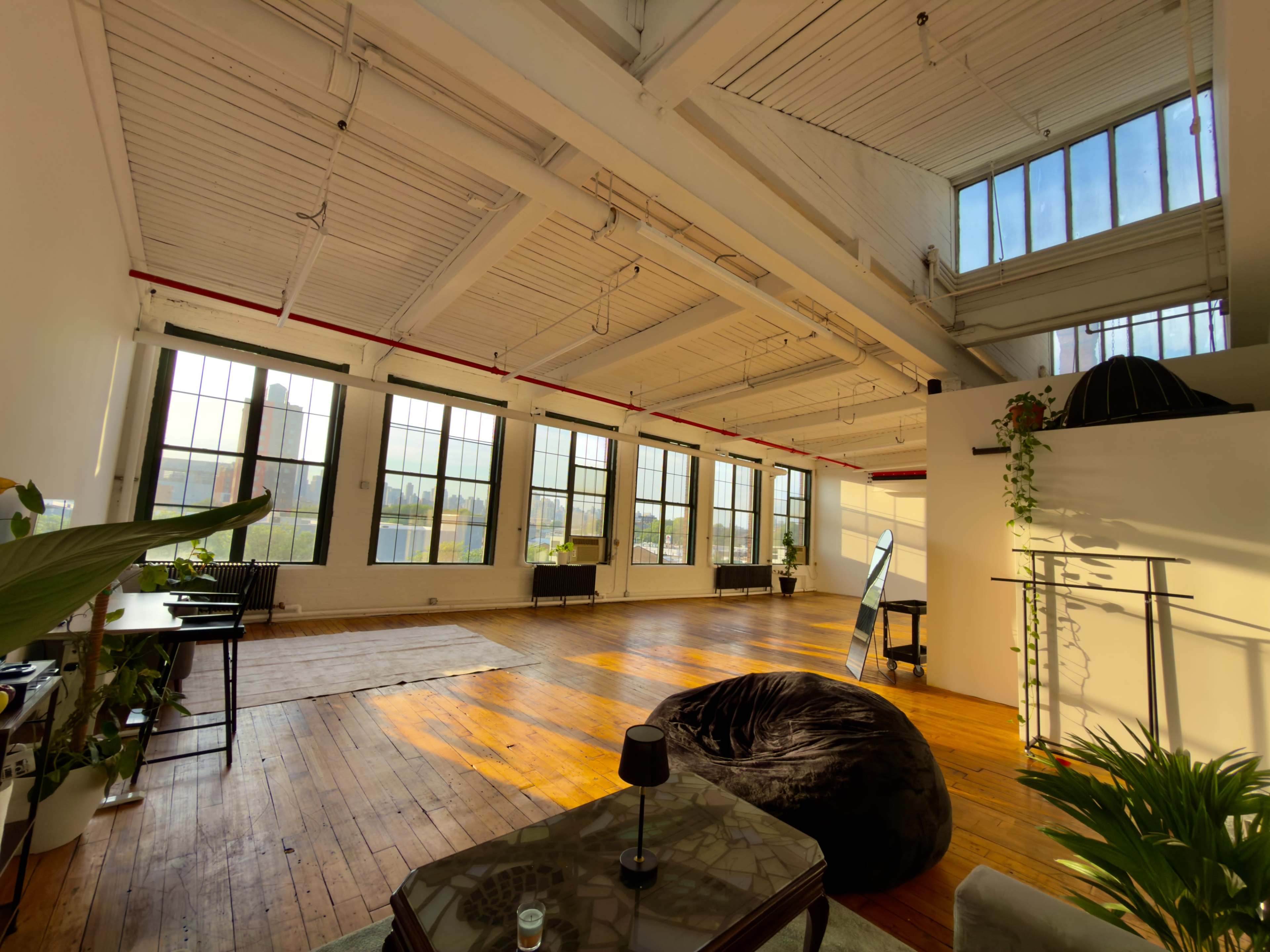The image shows a spacious, well-lit room with large windows, wooden flooring, and minimalist furniture arranged in an open layout.
