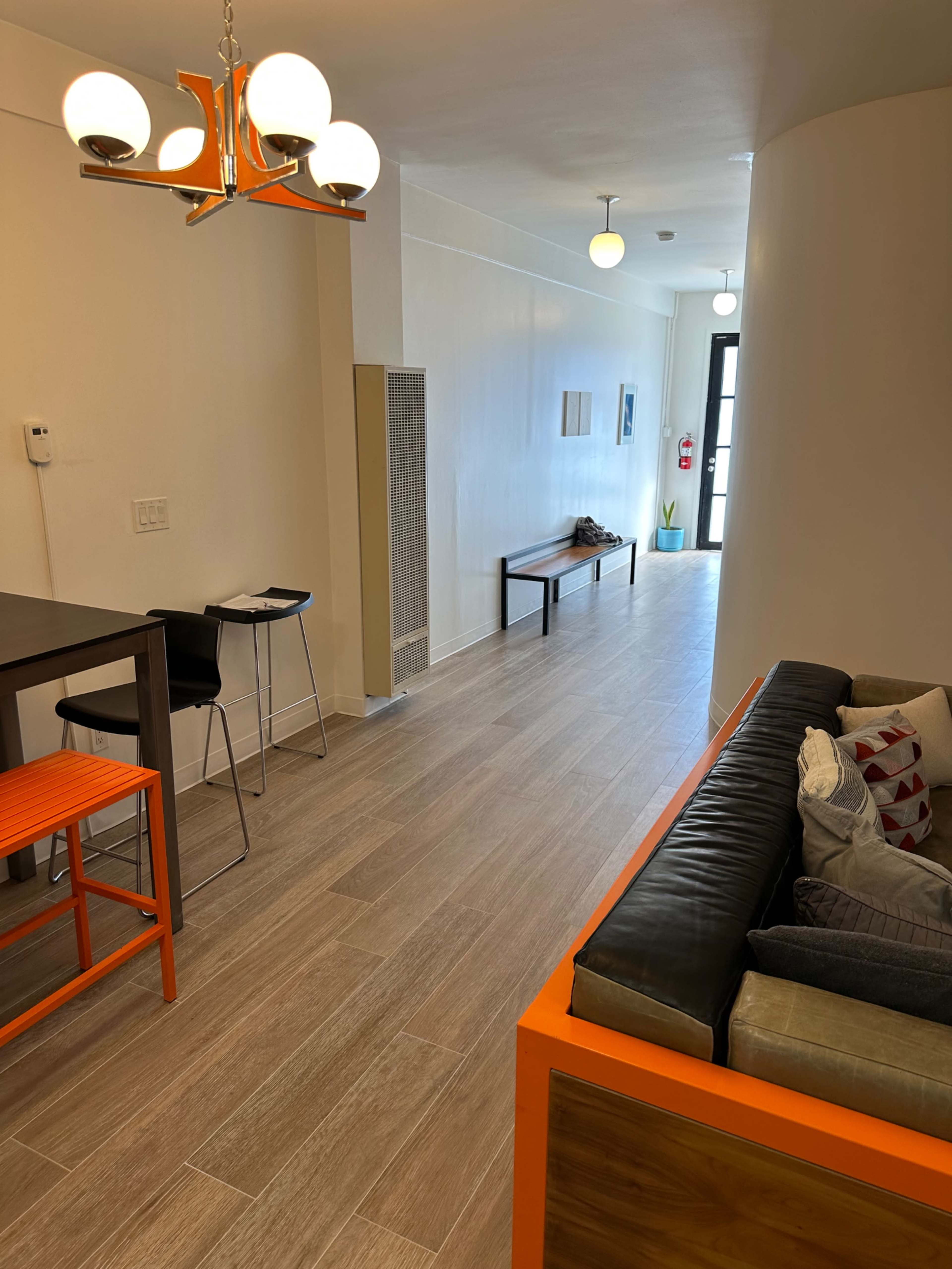 The image shows a modern interior space featuring a hallway with a table and chairs on one side, a sofa with decorative cushions on the other, and a glowing light fixture above.