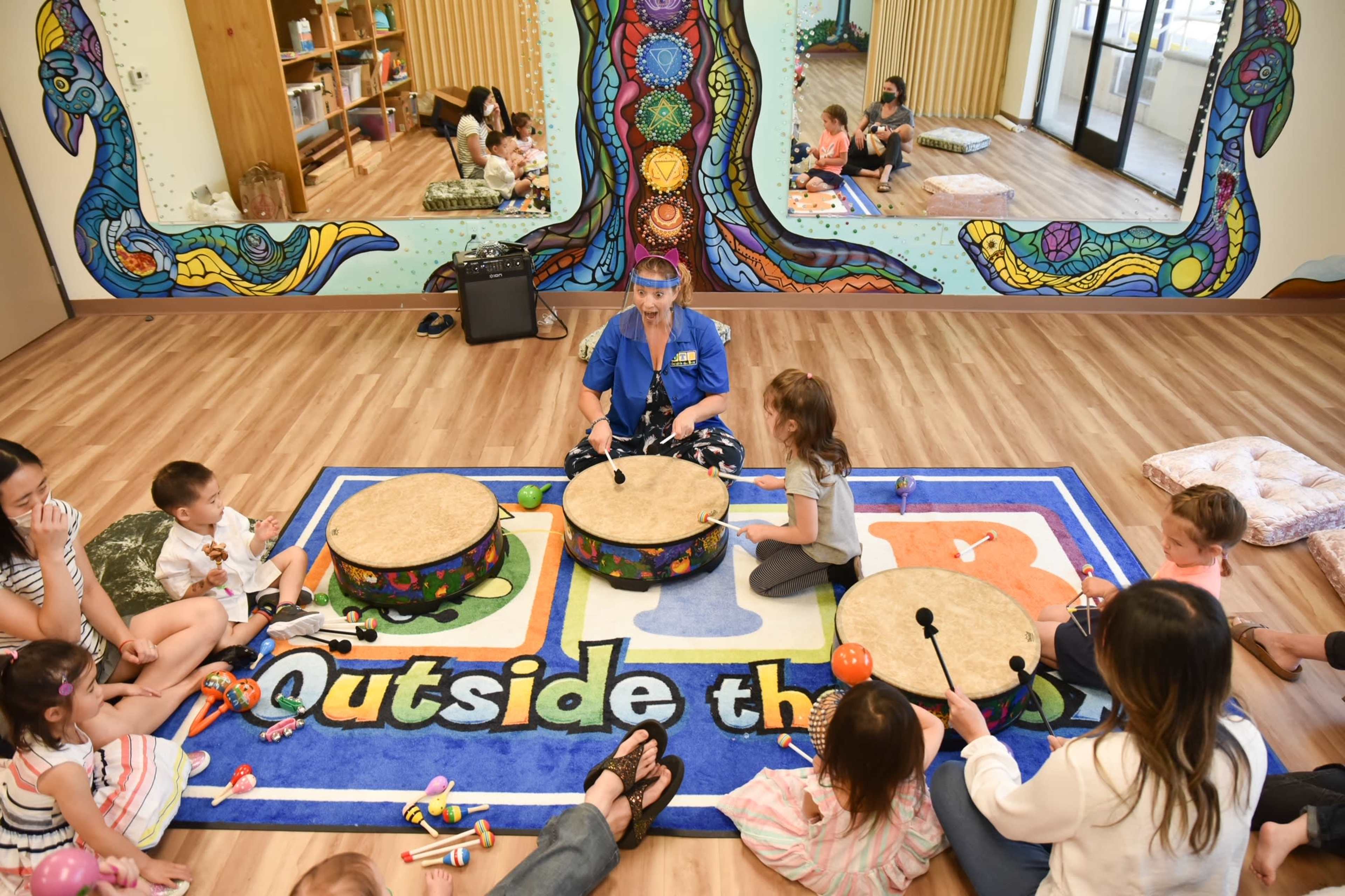 A group of children and an adult sit on the floor in a colorful classroom, engaging in a music session with various instruments.
