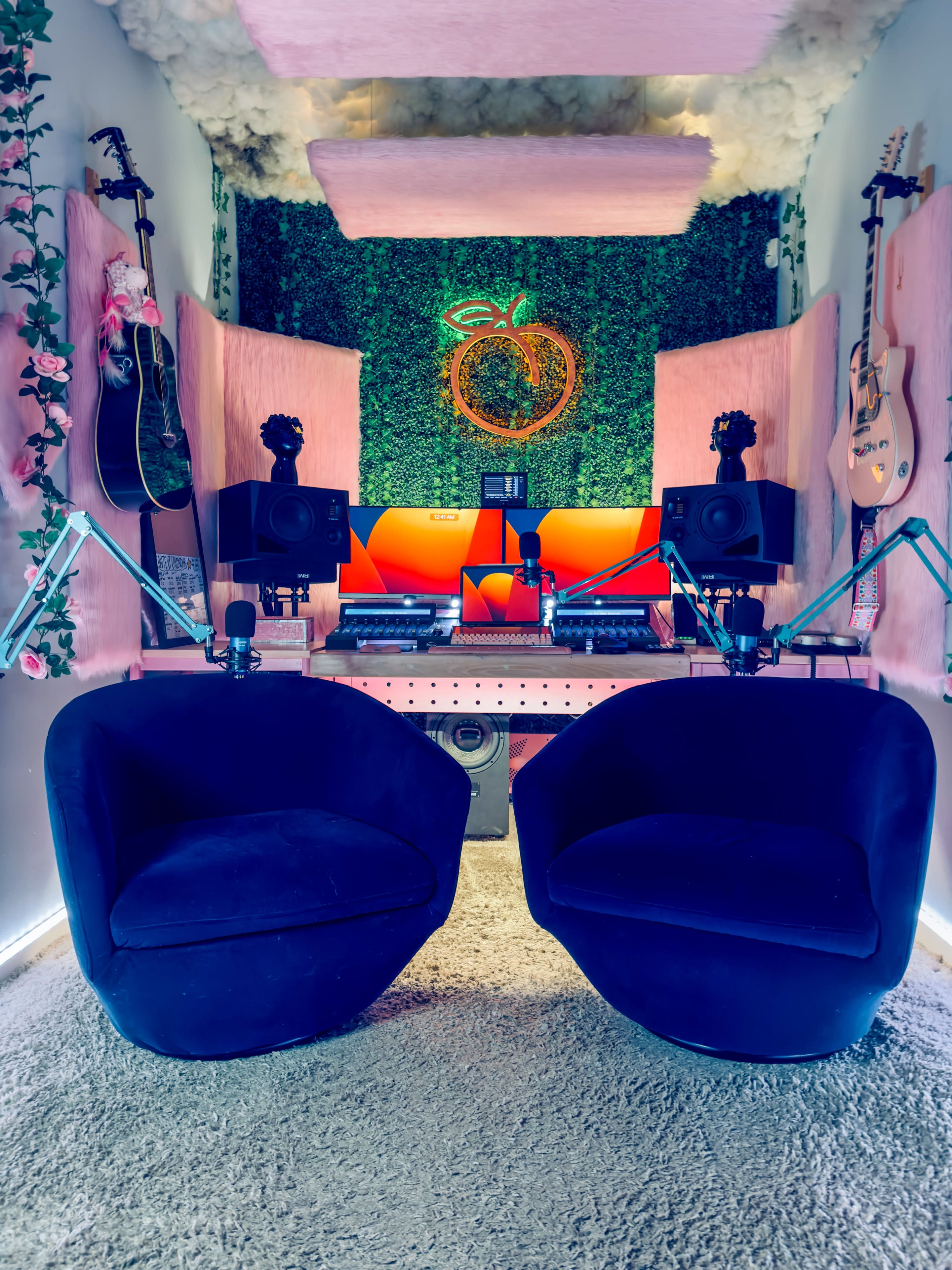 A small, cozy recording studio featuring two black chairs, musical instruments, sound equipment, and vibrant green wall decor with artificial foliage.