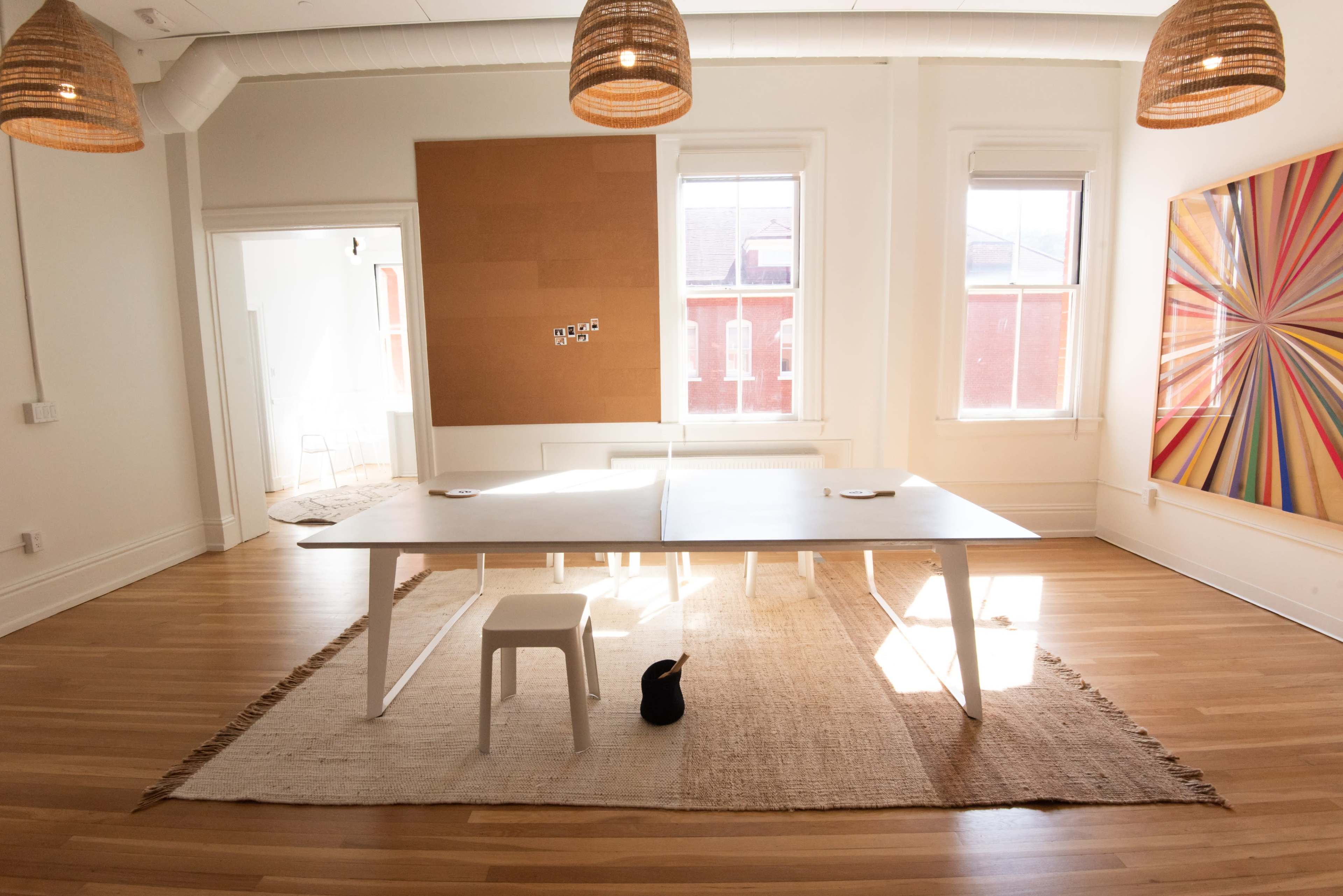 A spacious, well-lit room features a large white table with a black cat on a woven rug, surrounded by large windows and minimalist decor.