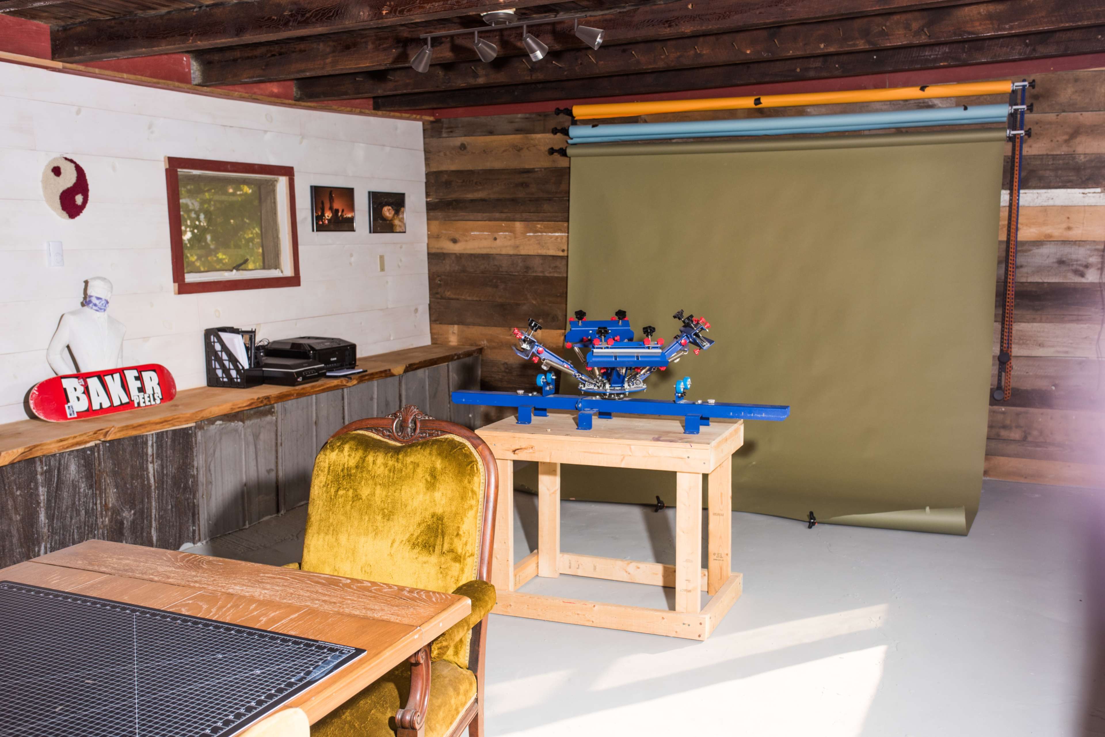 A wooden room with a green backdrop set up for photography, featuring a blue screen printing machine on a wooden table and a vintage chair nearby.