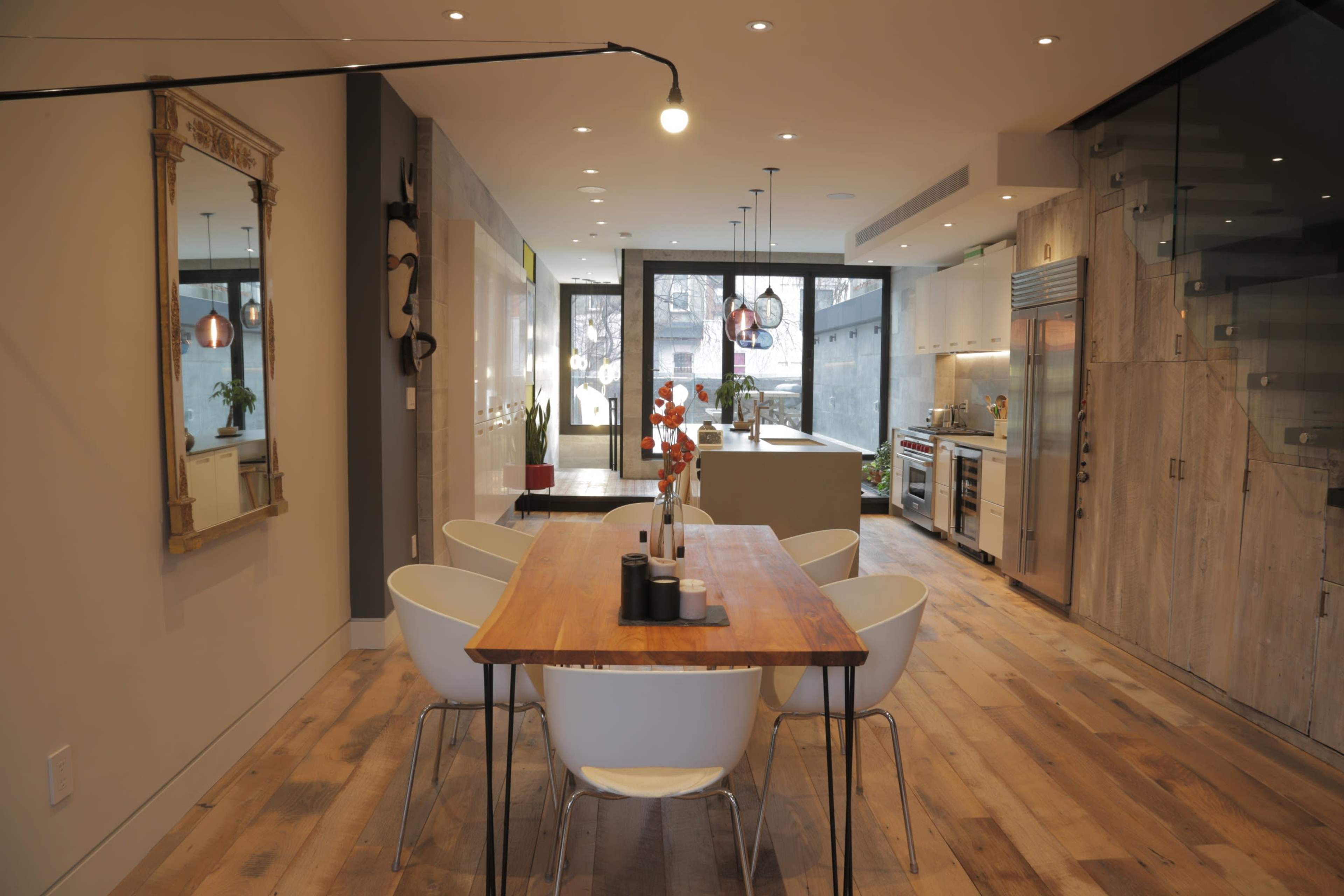 A spacious dining area features a wooden table surrounded by white chairs, leading into a modern kitchen with stainless steel appliances and a large window.