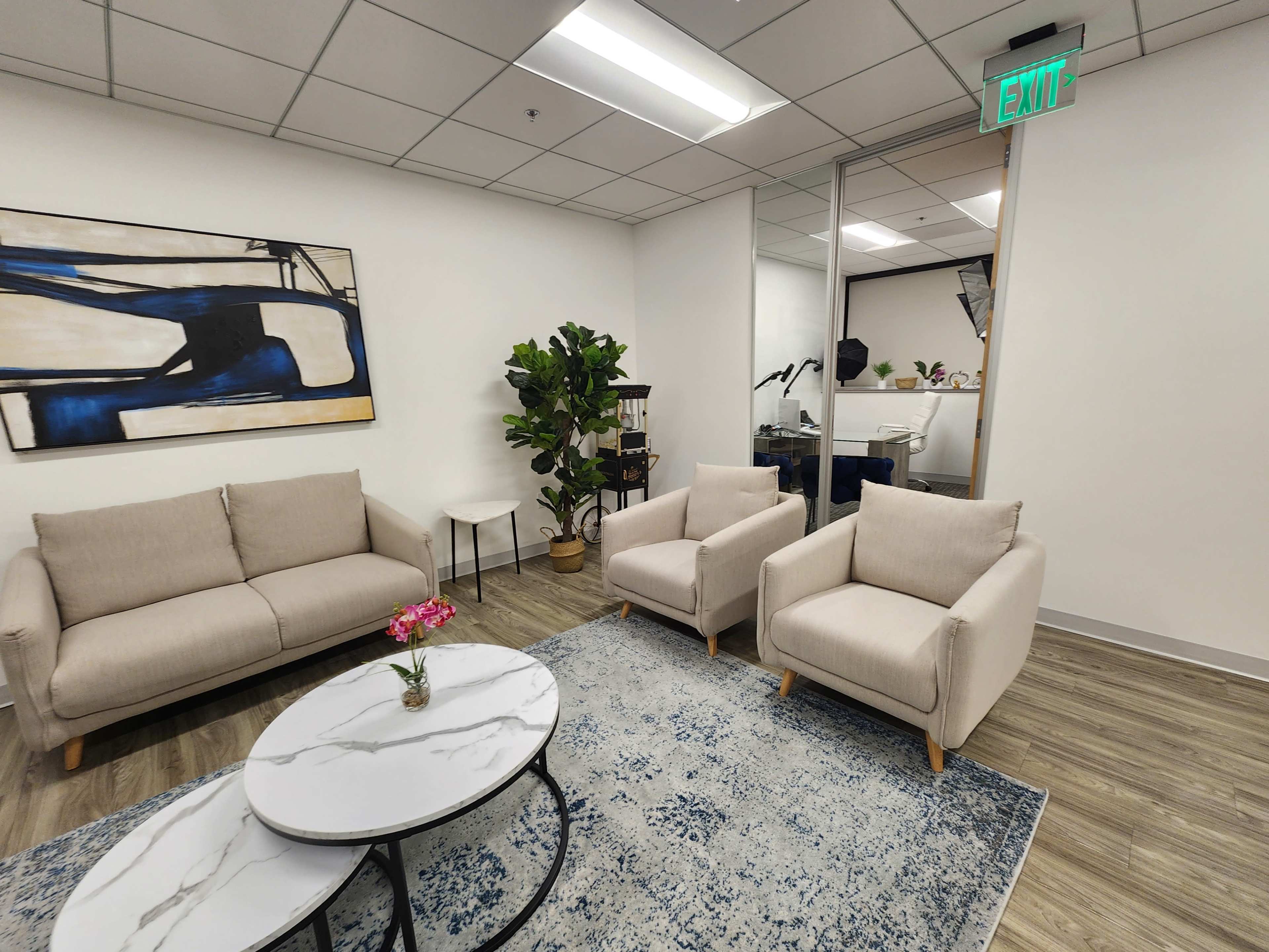 The image shows a modern office lounge area with two beige chairs, a gray and white marble coffee table, a small side table, and a painting on the wall, alongside a potted plant.