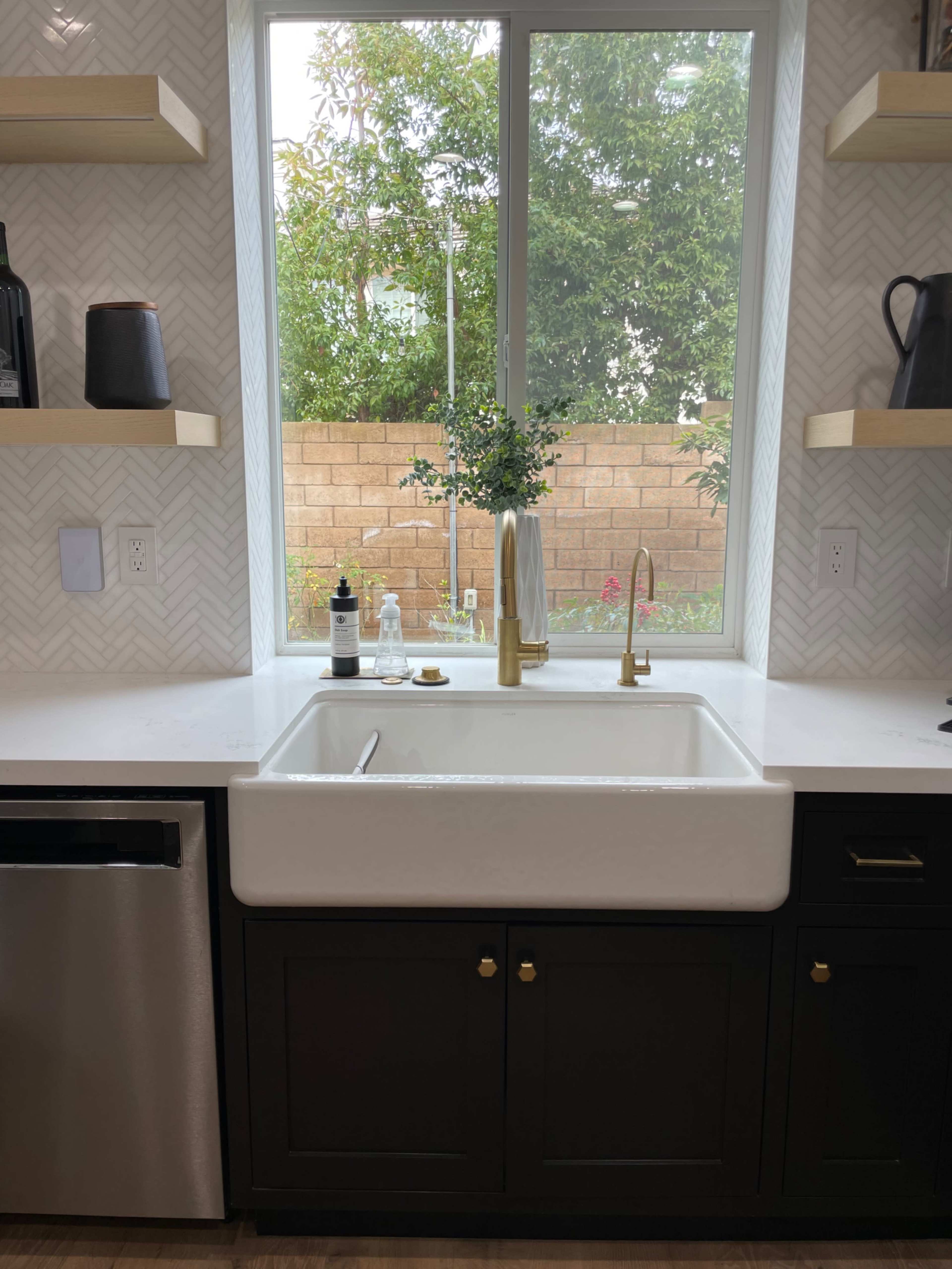 A white farmhouse sink is positioned beneath a window with a view of a garden, surrounded by dark cabinetry and shelving.
