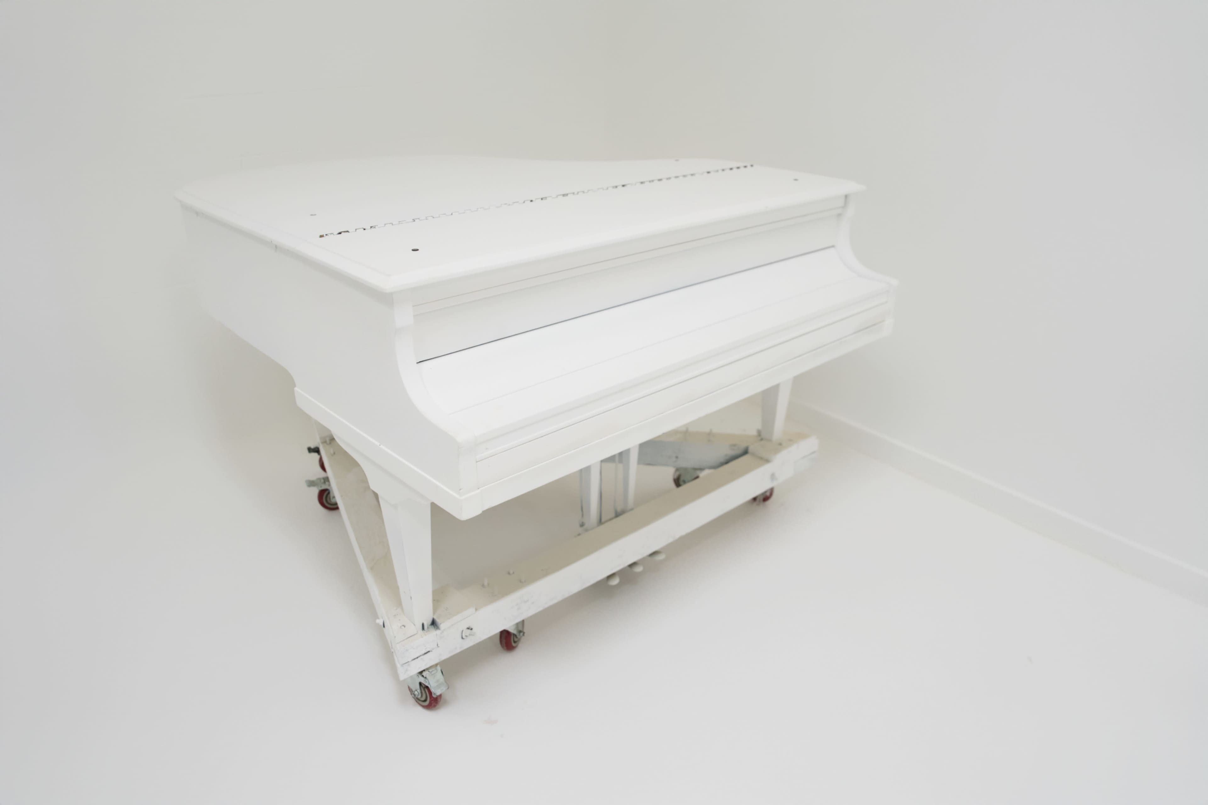 A white grand piano is positioned on wheels, set against a plain white background.