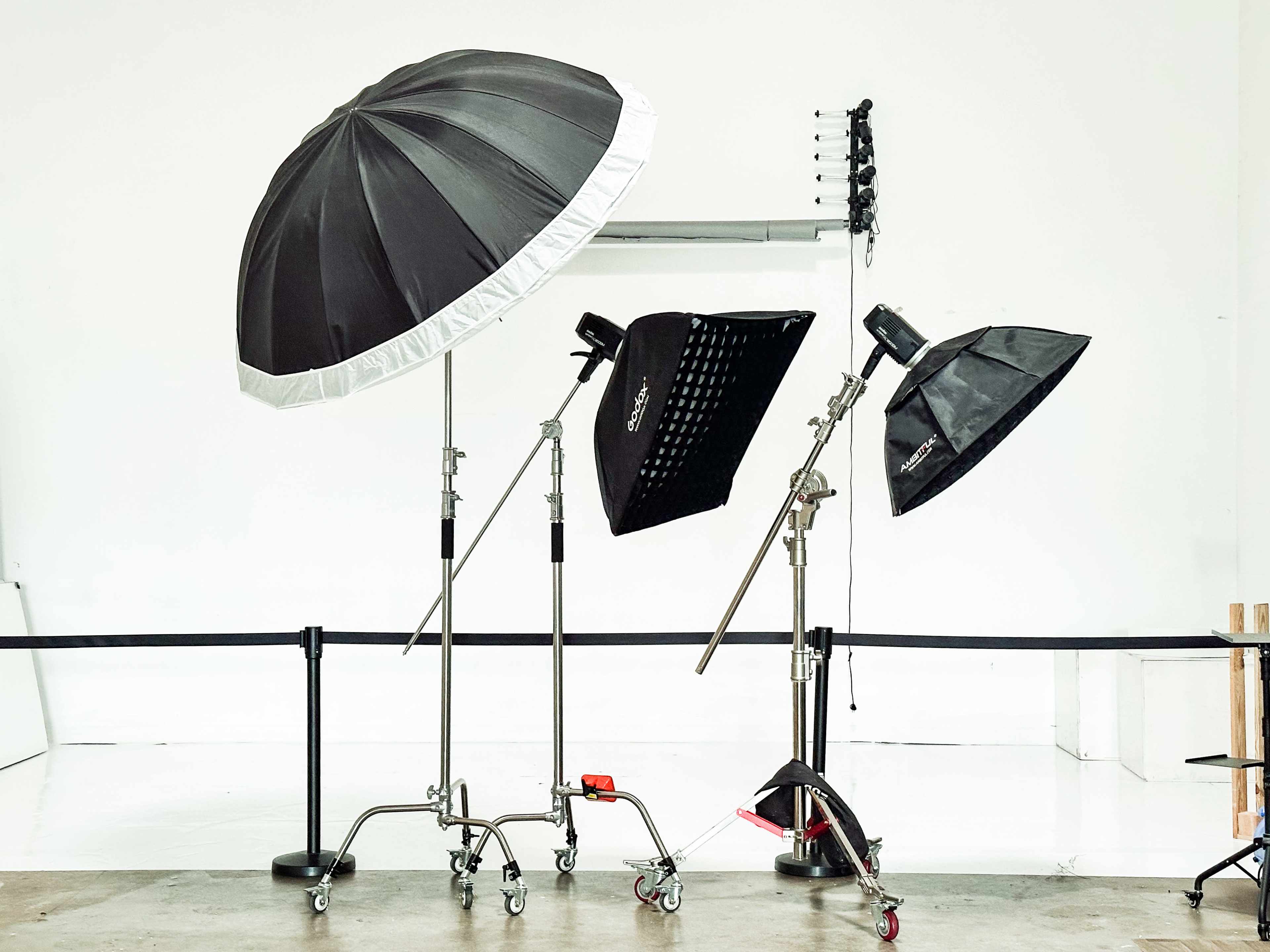 The image shows various photography lighting equipment, including umbrellas, softboxes, and light stands, arranged in a studio setting.