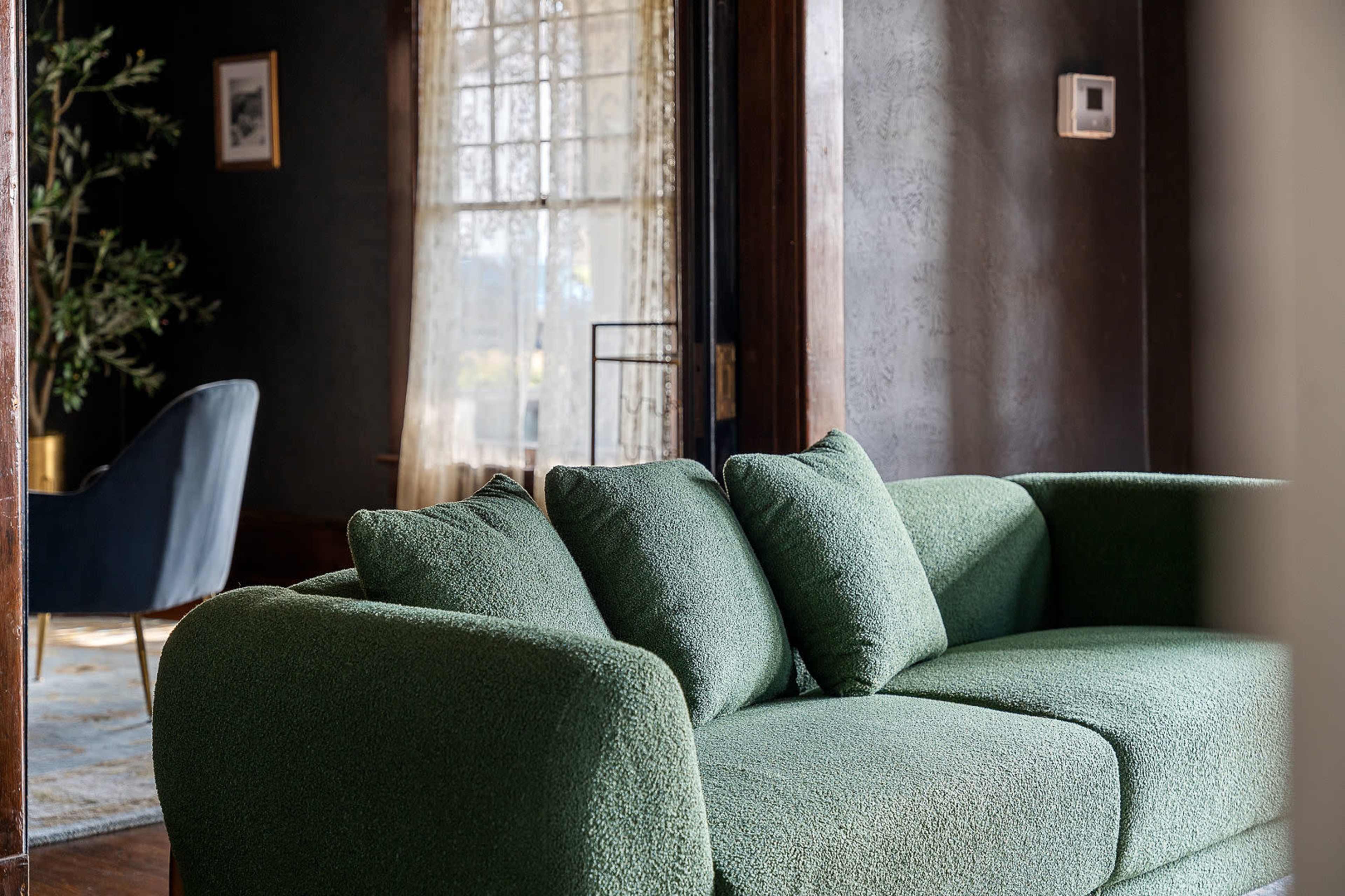 The image shows a green sofa with multiple cushions in a room featuring dark walls and a window with sheer curtains in the background.
