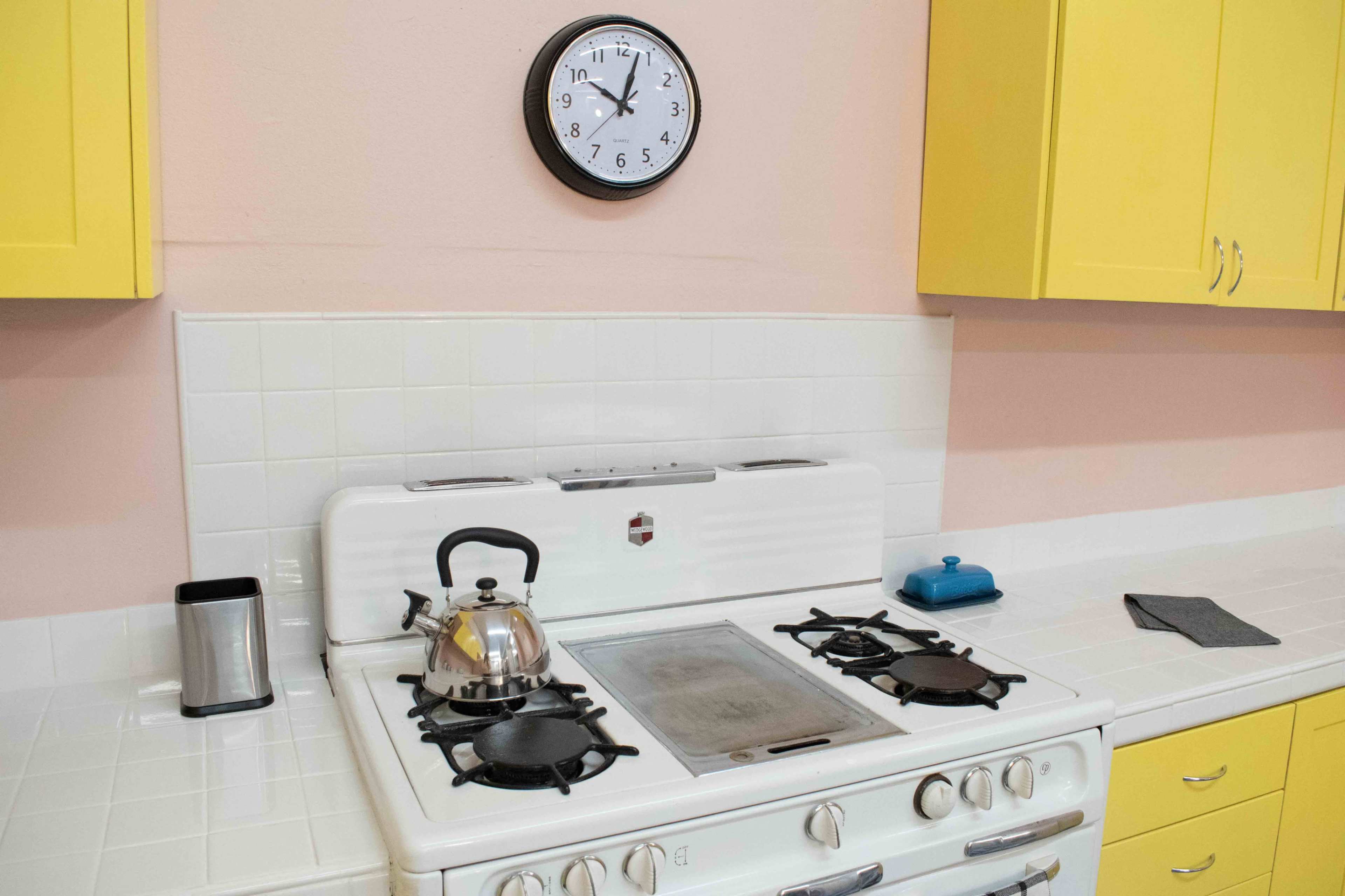A white gas stove with a kettle on top is situated under a wall clock and surrounded by yellow cabinets in a kitchen.