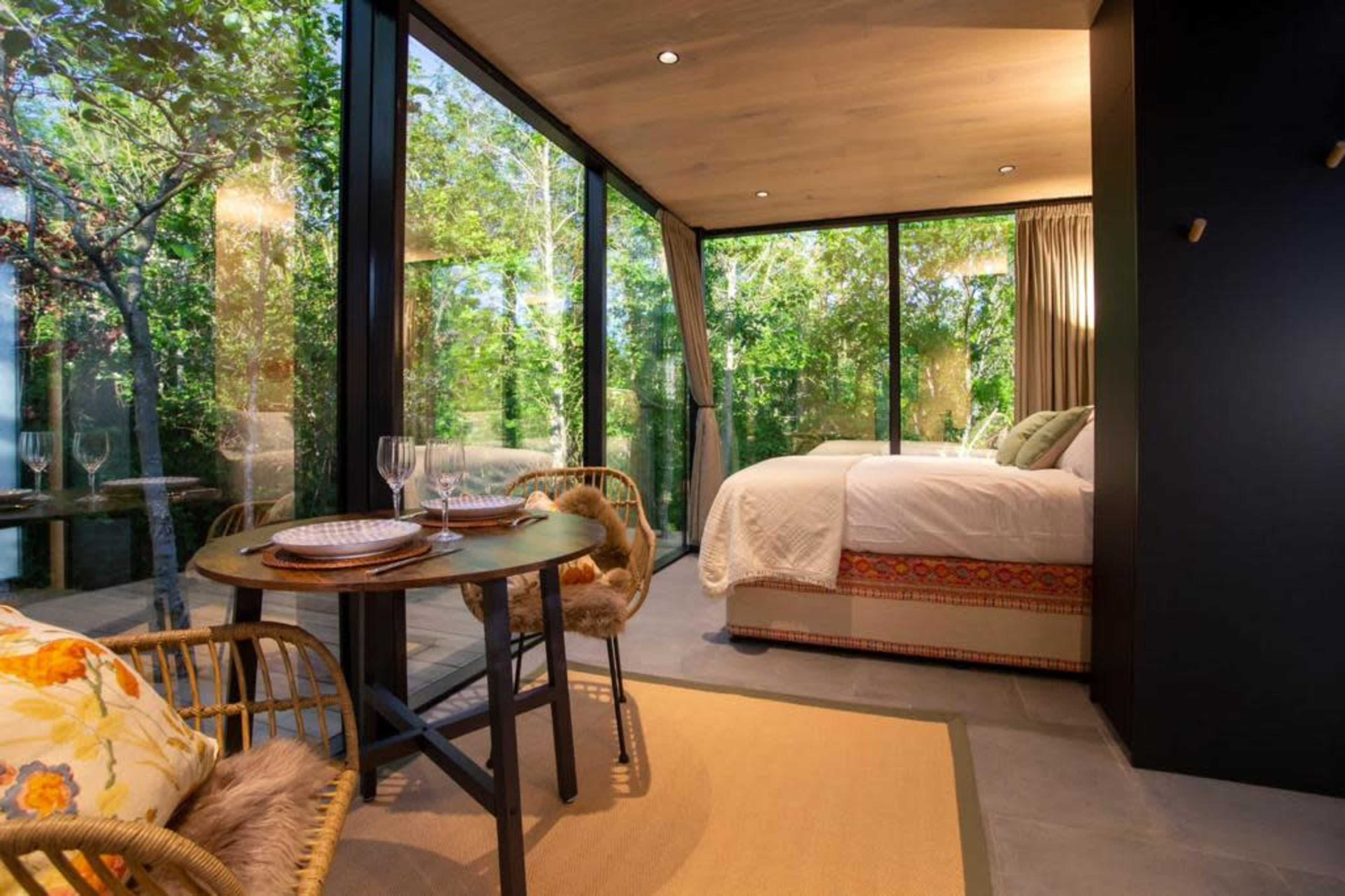 A cozy bedroom with a large glass wall overlooking a lush green forest features a dining table set for two.