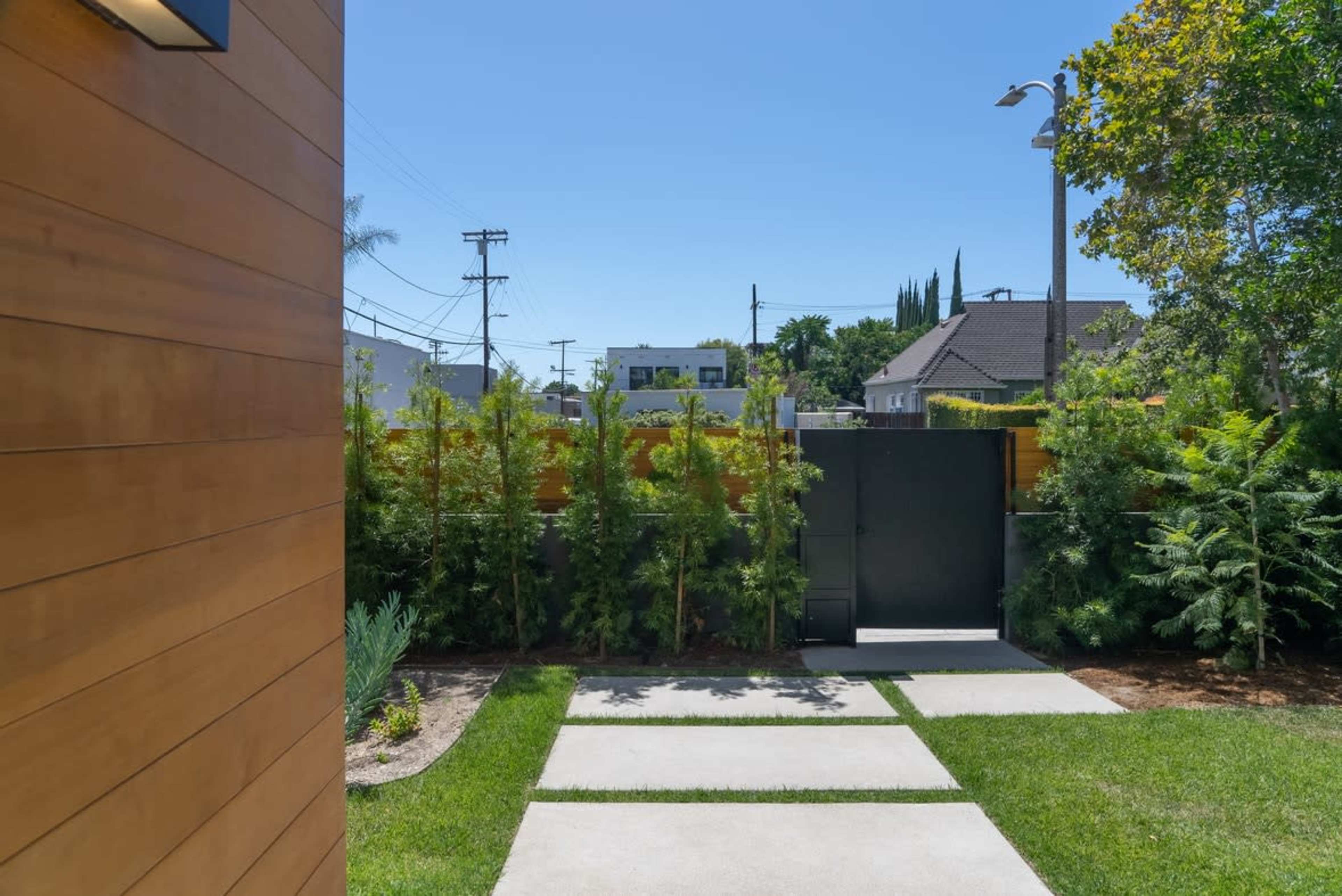 A pathway leading to a black gate, flanked by lush greenery and surrounded by residential buildings under a clear blue sky.