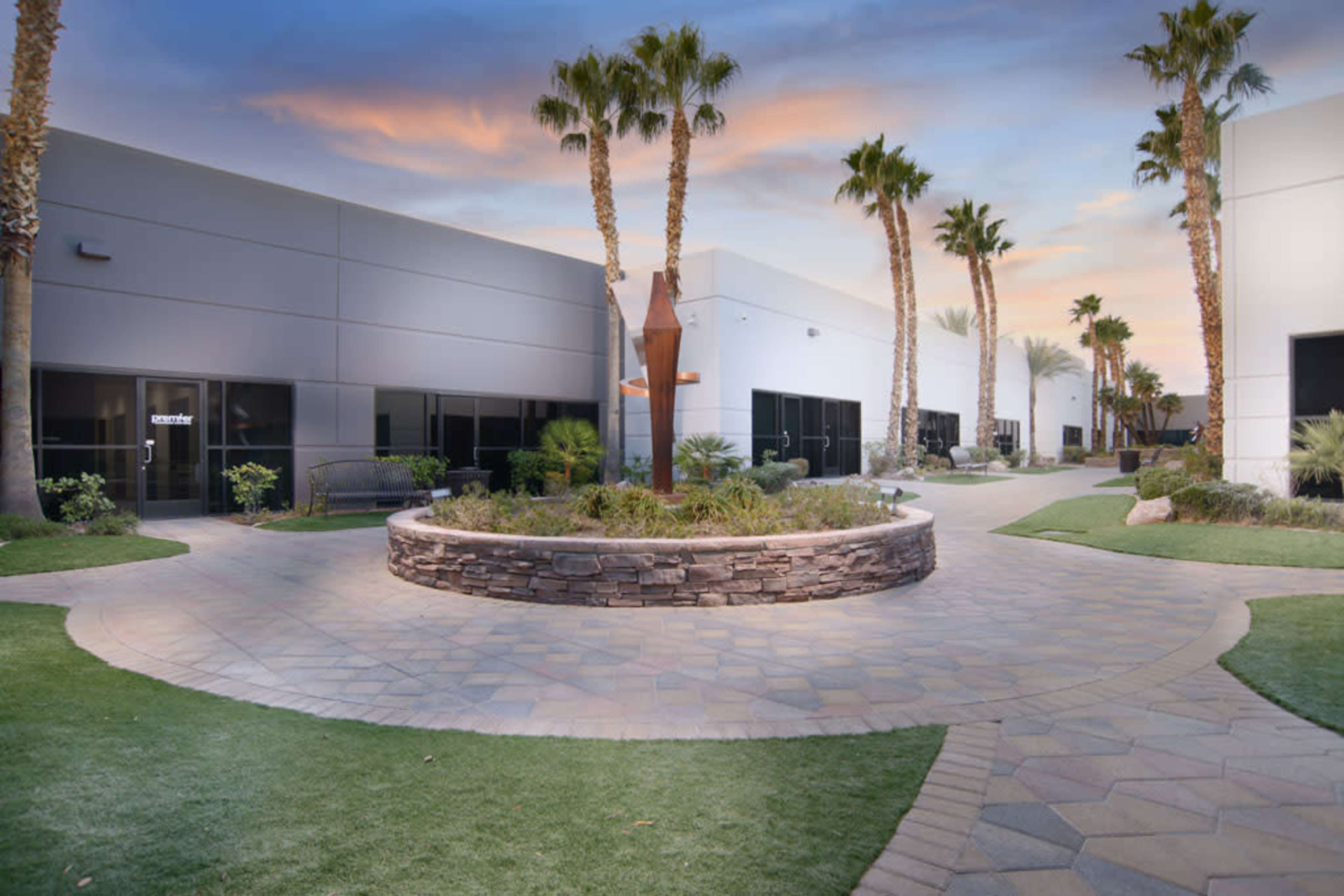 The image shows a curved pathway surrounded by palm trees and modern buildings, leading to a stone planter in a landscaped area at sunset.