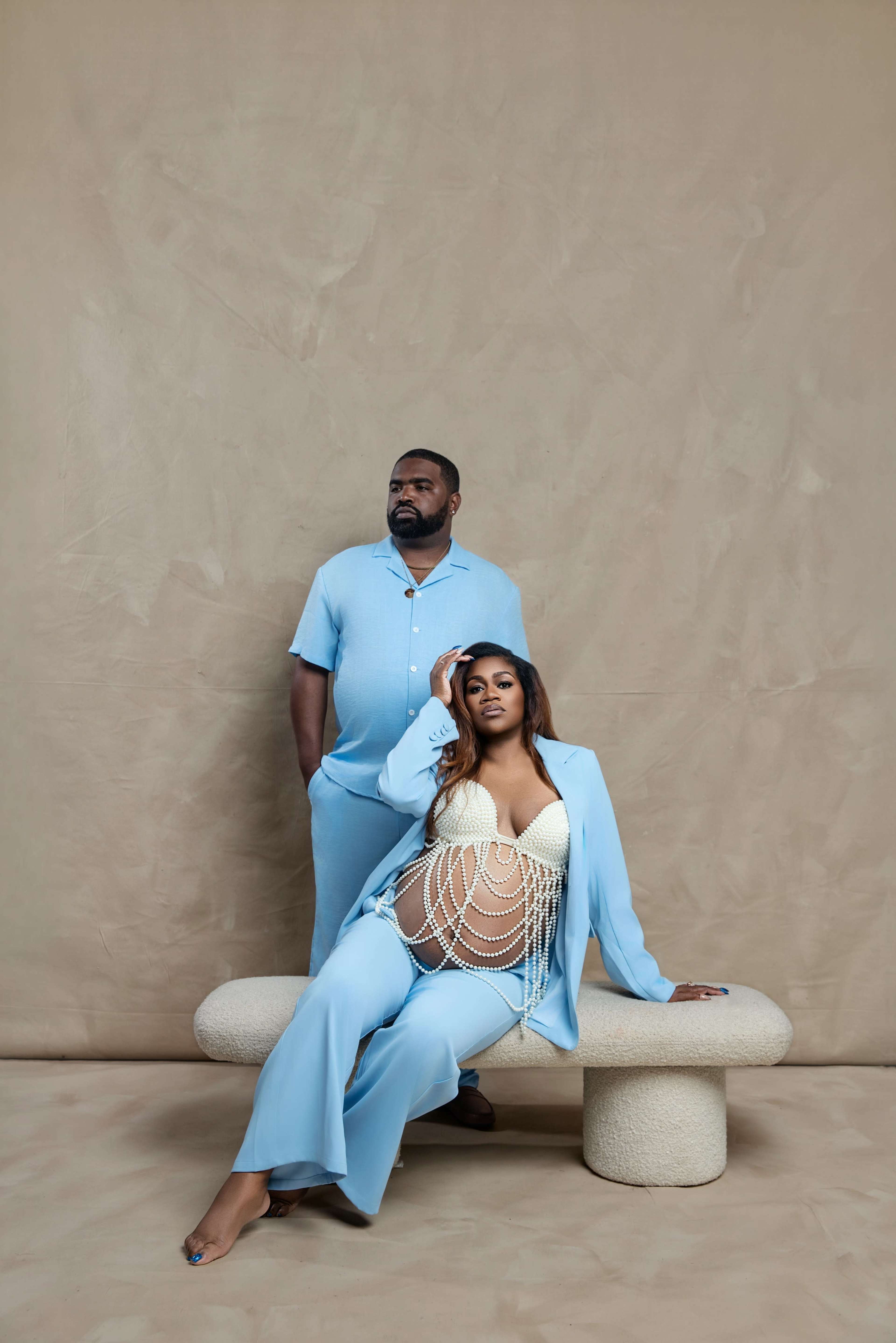 A pregnant woman in a pearl-adorned outfit sits on a stone-like structure, while a man stands behind her, both wearing matching light blue clothing.