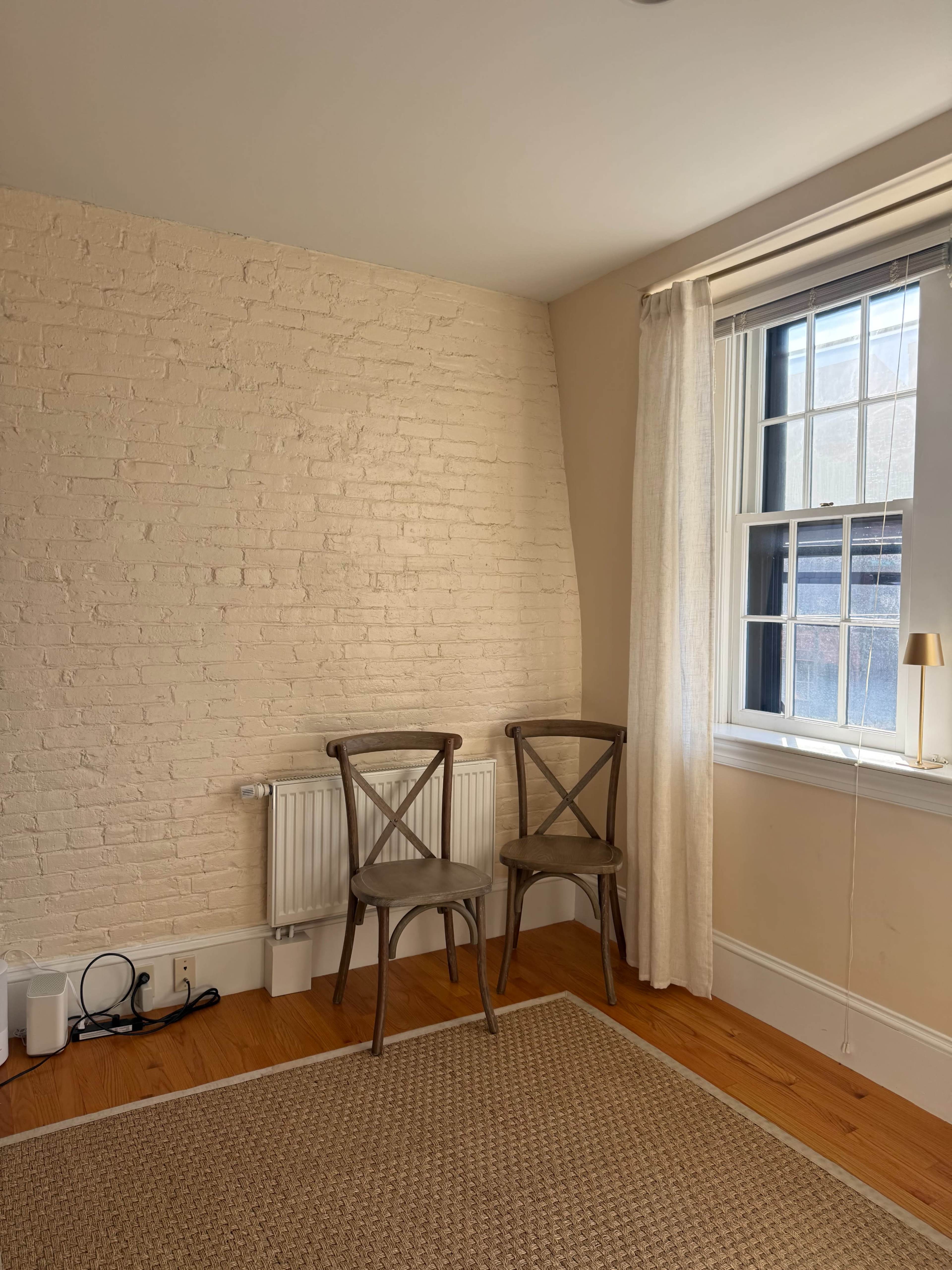 The image shows a corner of a room with two wooden chairs next to a radiator, a large window with curtains, and a woven rug on a hardwood floor.