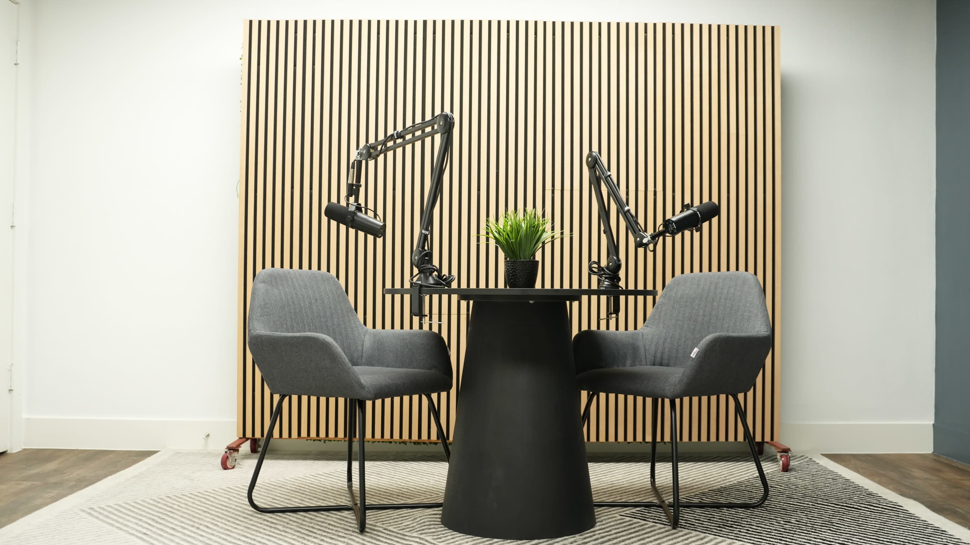 A round black table is positioned between two gray chairs in front of a wooden slat wall with two adjustable microphone arms mounted on it.