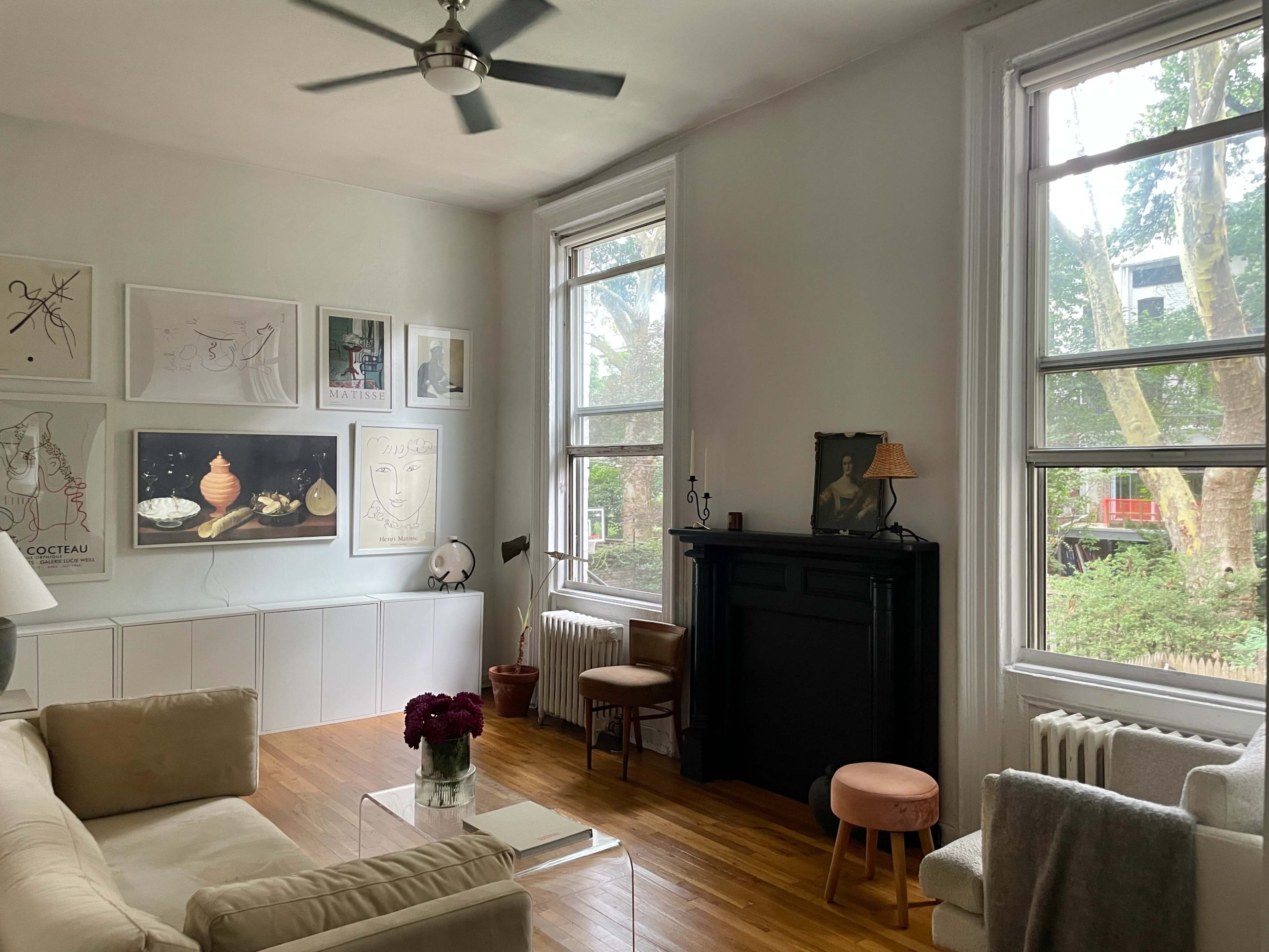 The room features a light-colored sofa, a glass coffee table, and a decorative fireplace, with large windows allowing natural light and views of greenery outside.