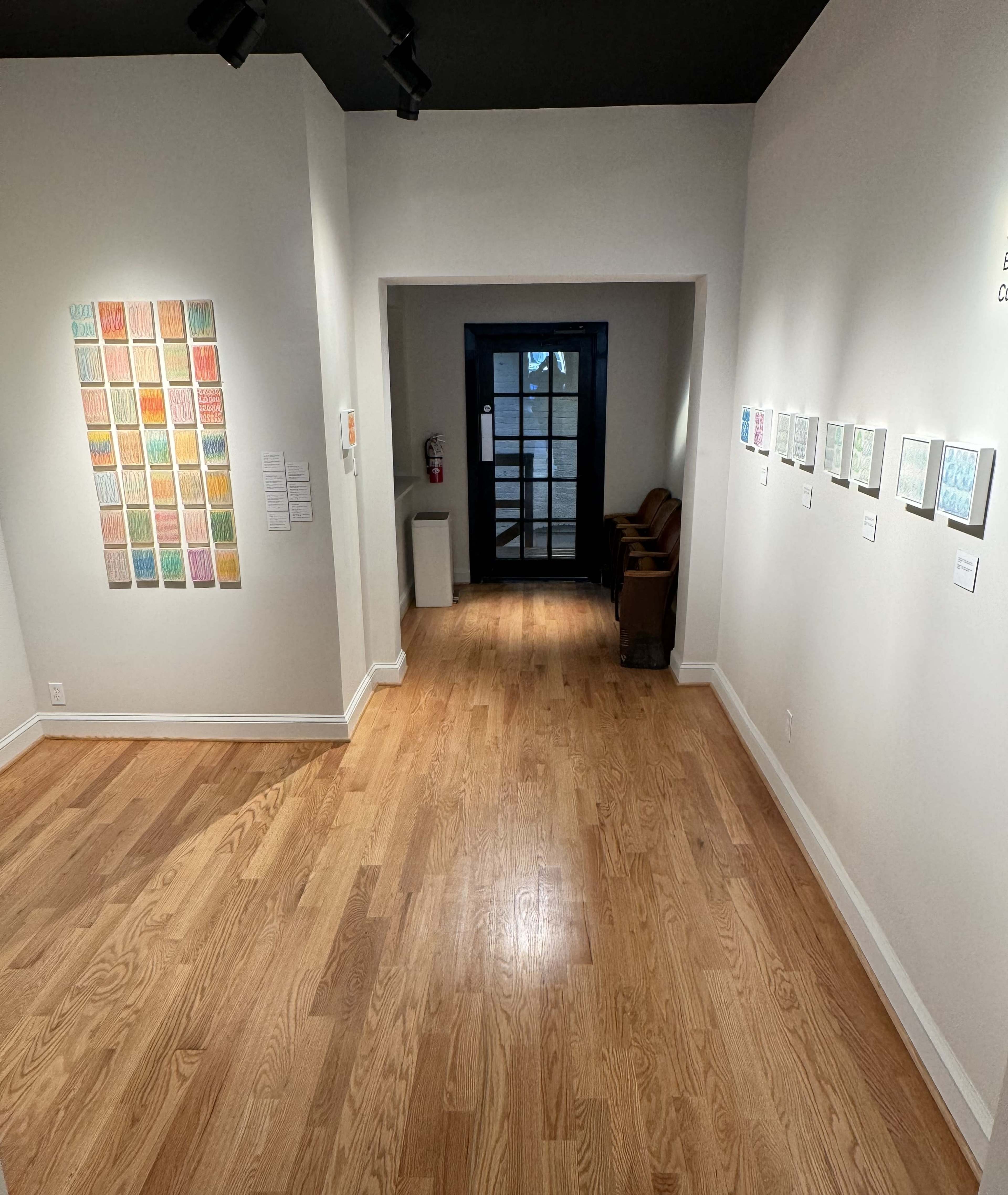 An art gallery with wooden flooring, featuring colorful art pieces displayed on the walls and a doorway leading to another room.