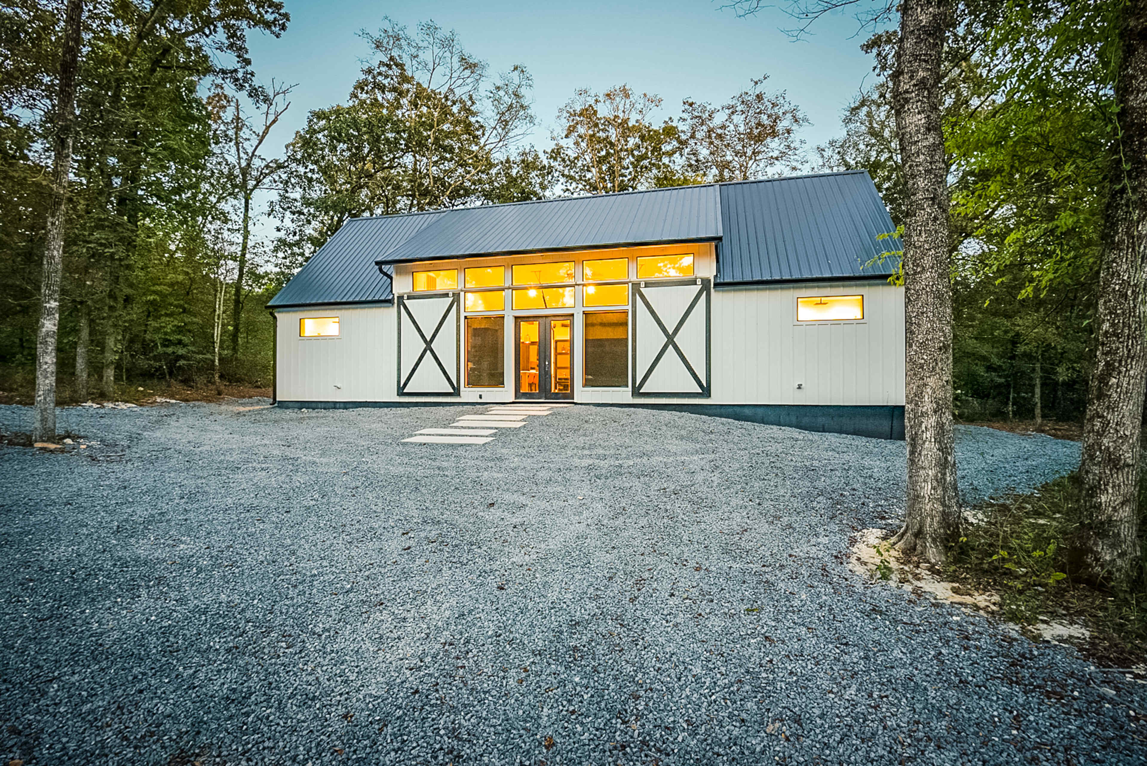 A modern, single-story house with a metal roof and large front windows is surrounded by a gravel driveway and trees.