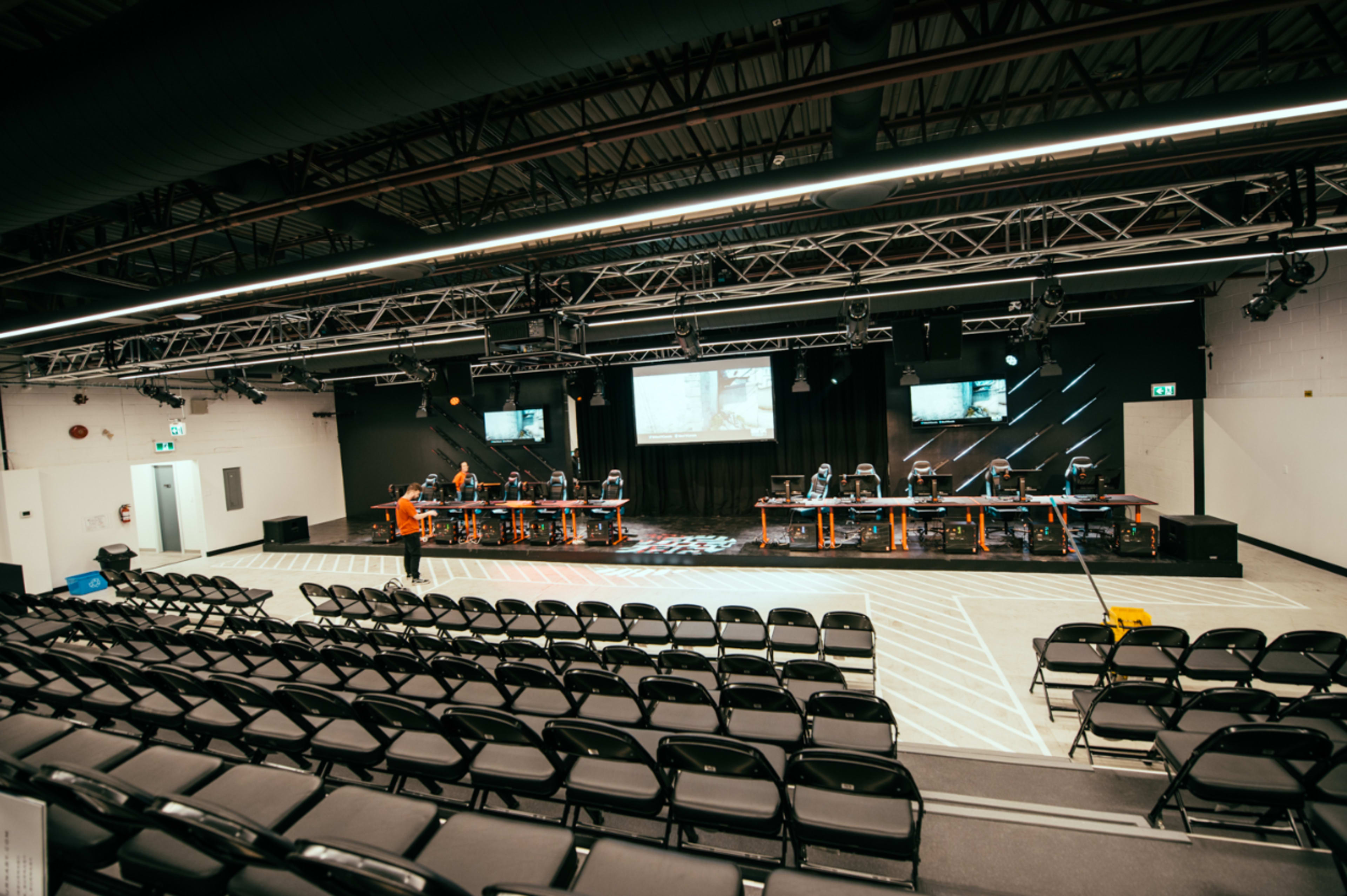 An empty auditorium features rows of black chairs facing a stage with multiple computer stations and screens.