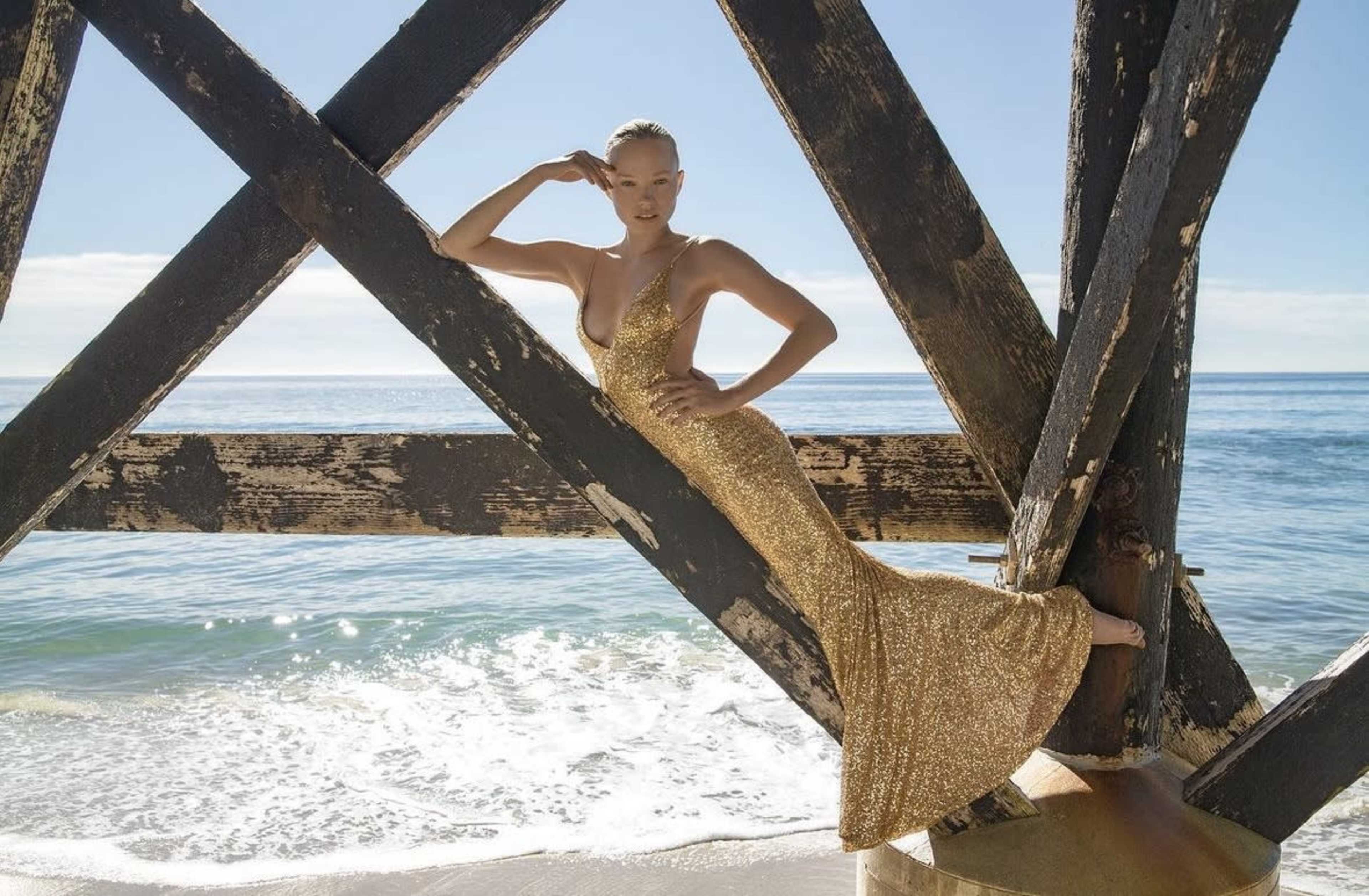 A woman in a gold sequined dress poses against a weathered wooden pier by the beach with waves rolling in.