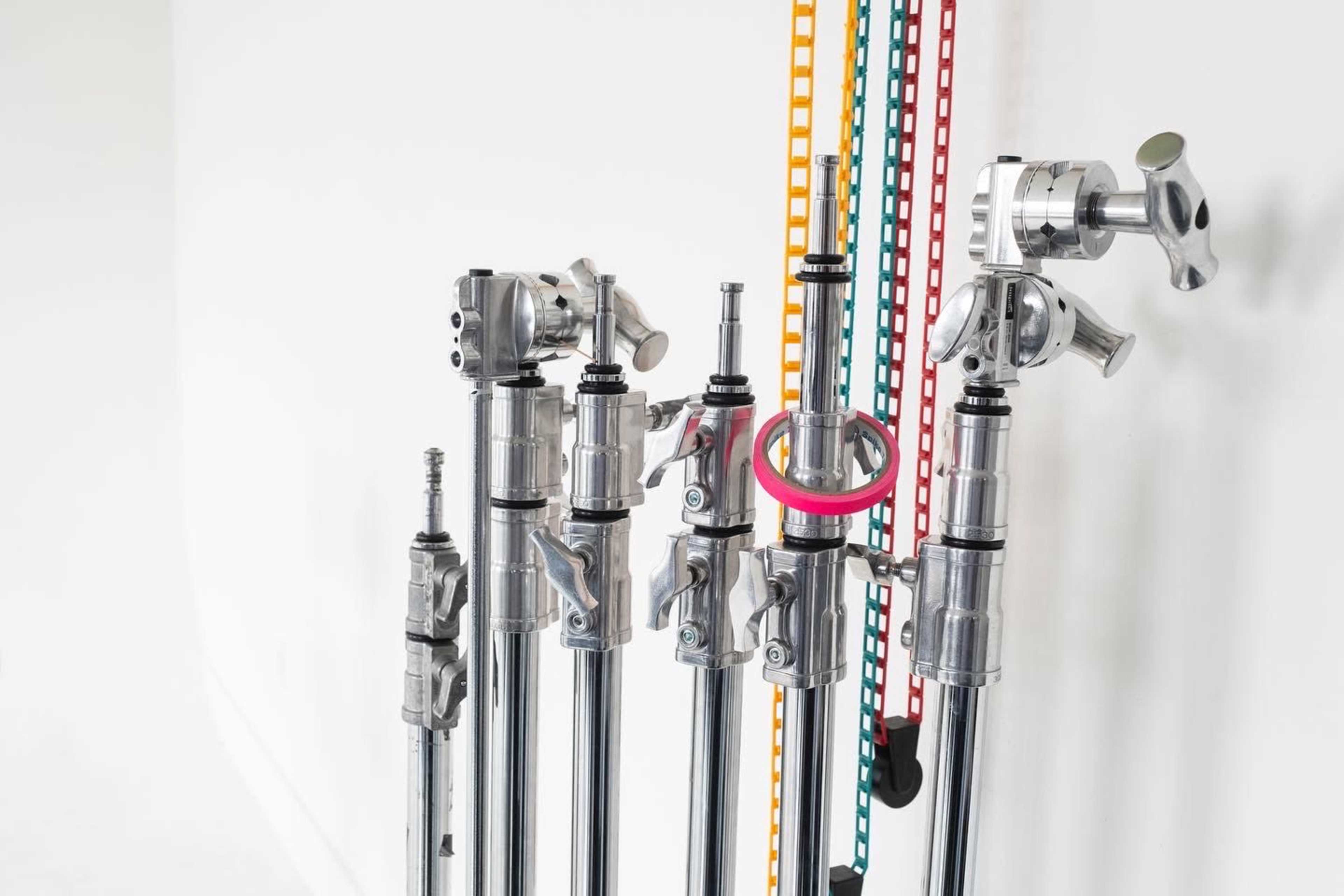 The image shows a collection of photo or video support stands lined up against a white wall, with colorful straps hanging nearby.
