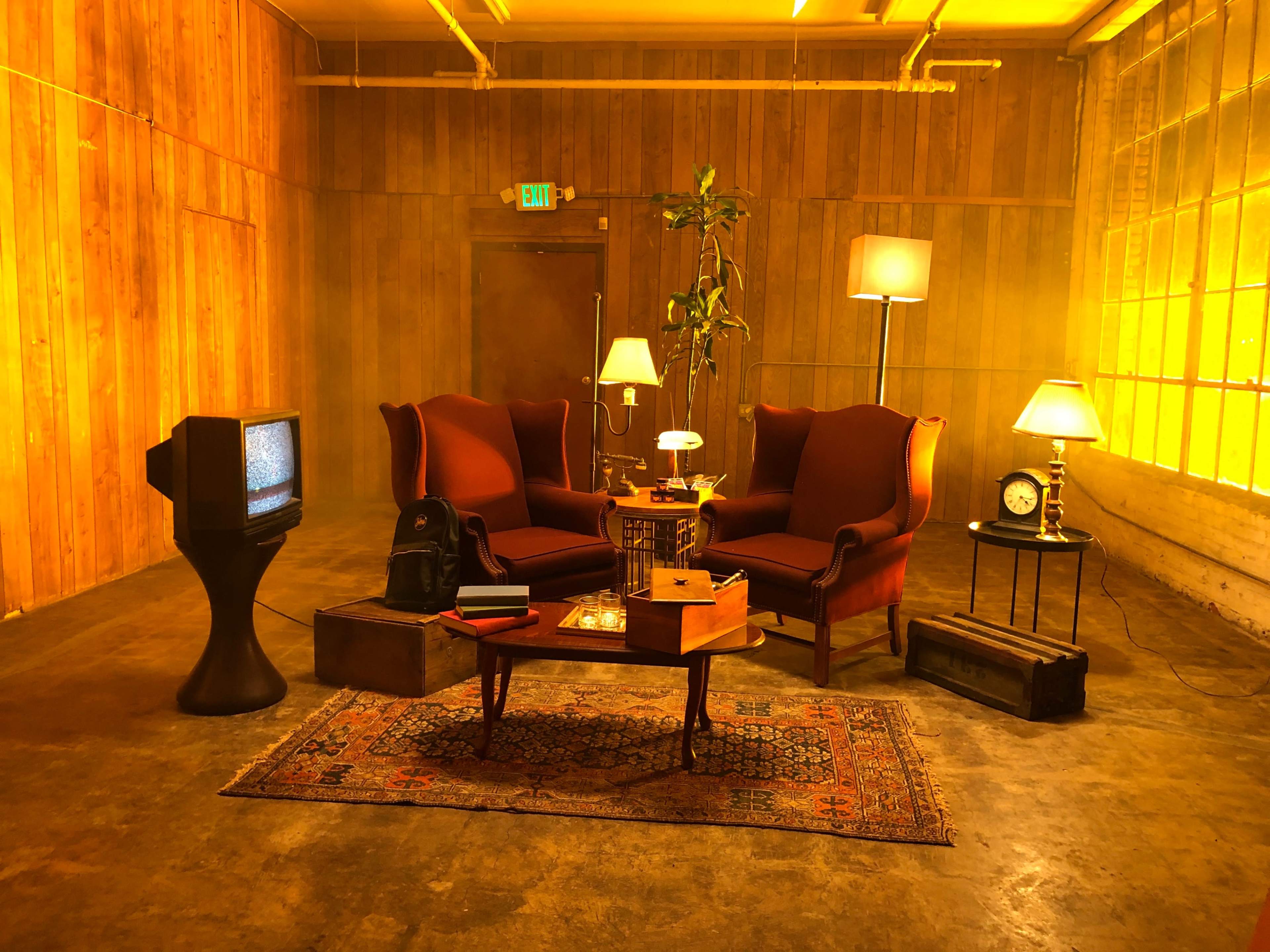 The scene features two red armchairs facing each other in a room with wooden walls, illuminated by warm lighting, accompanied by a vintage television, a side table with a lamp, a clock, and a potted plant.