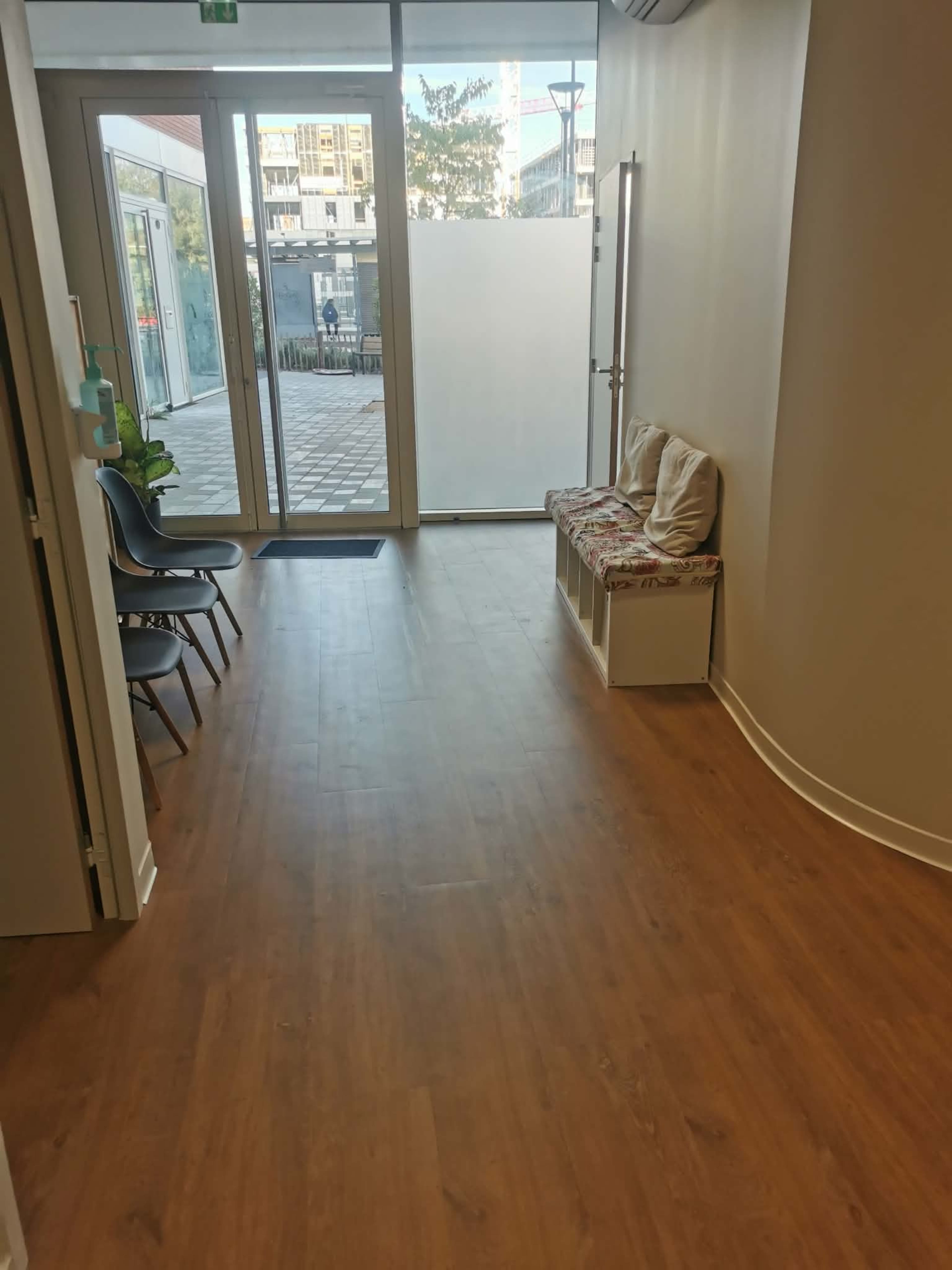 The image shows a hallway with a wooden floor, seating along one side, and large glass doors leading to an outdoor area.